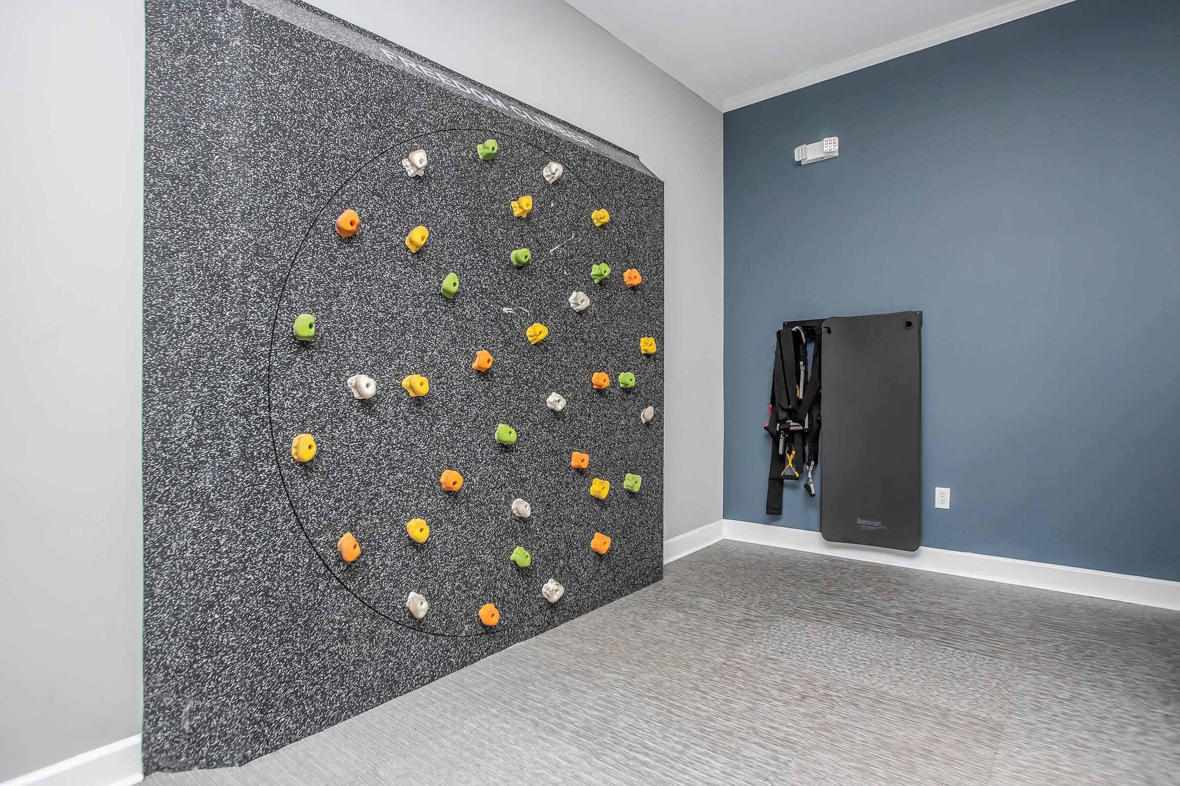A brightly colored climbing wall with various holds in orange, green, yellow, and white, set against a grey textured background. A crash pad is positioned to the right, and the wall is located in a room with blue walls and carpeted flooring.