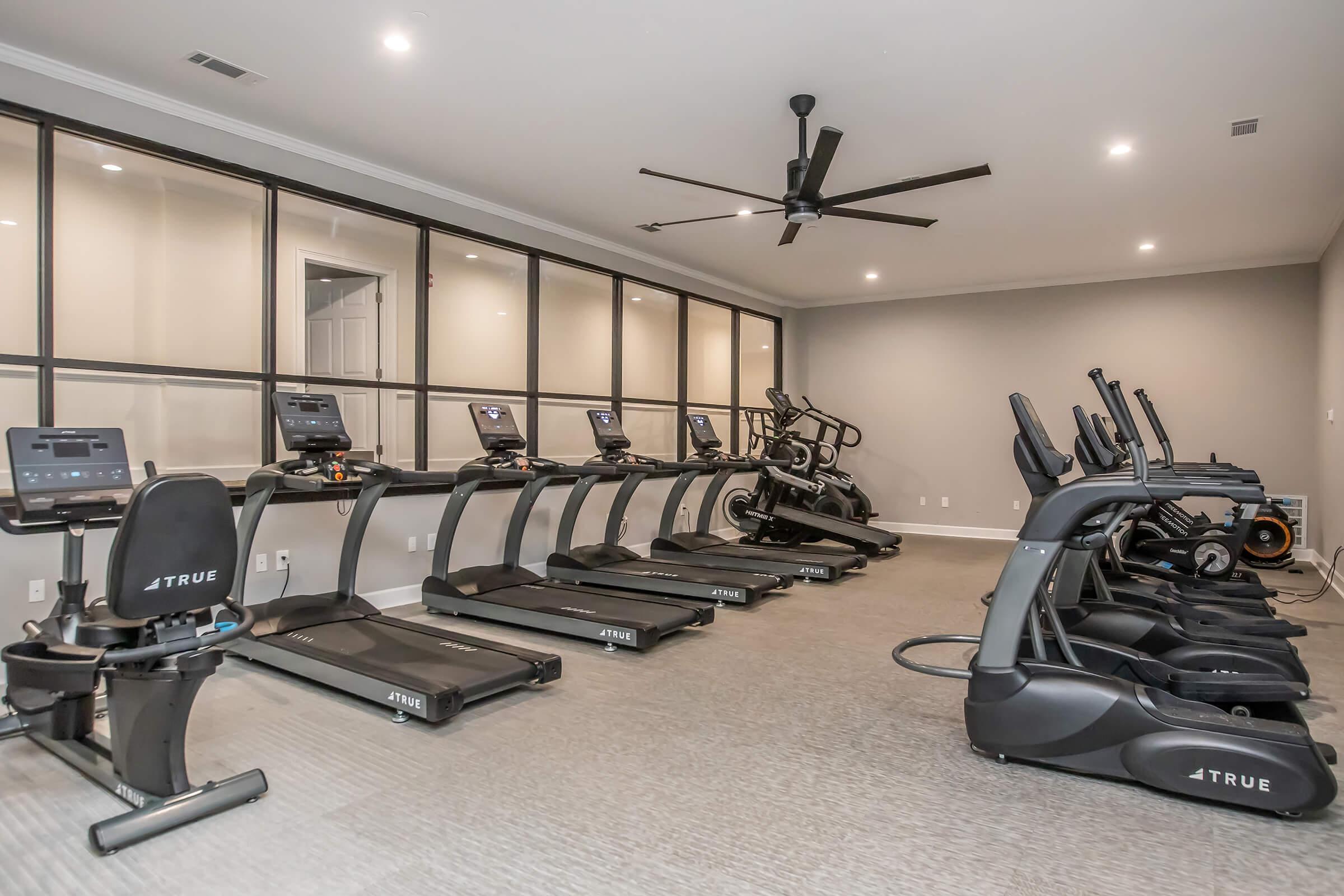 A modern gym featuring multiple treadmills and elliptical machines arranged in a spacious room with a neutral-colored wall, a ceiling fan, and ample lighting. The workout area is designed for fitness enthusiasts, providing a clean and well-organized environment for exercise.