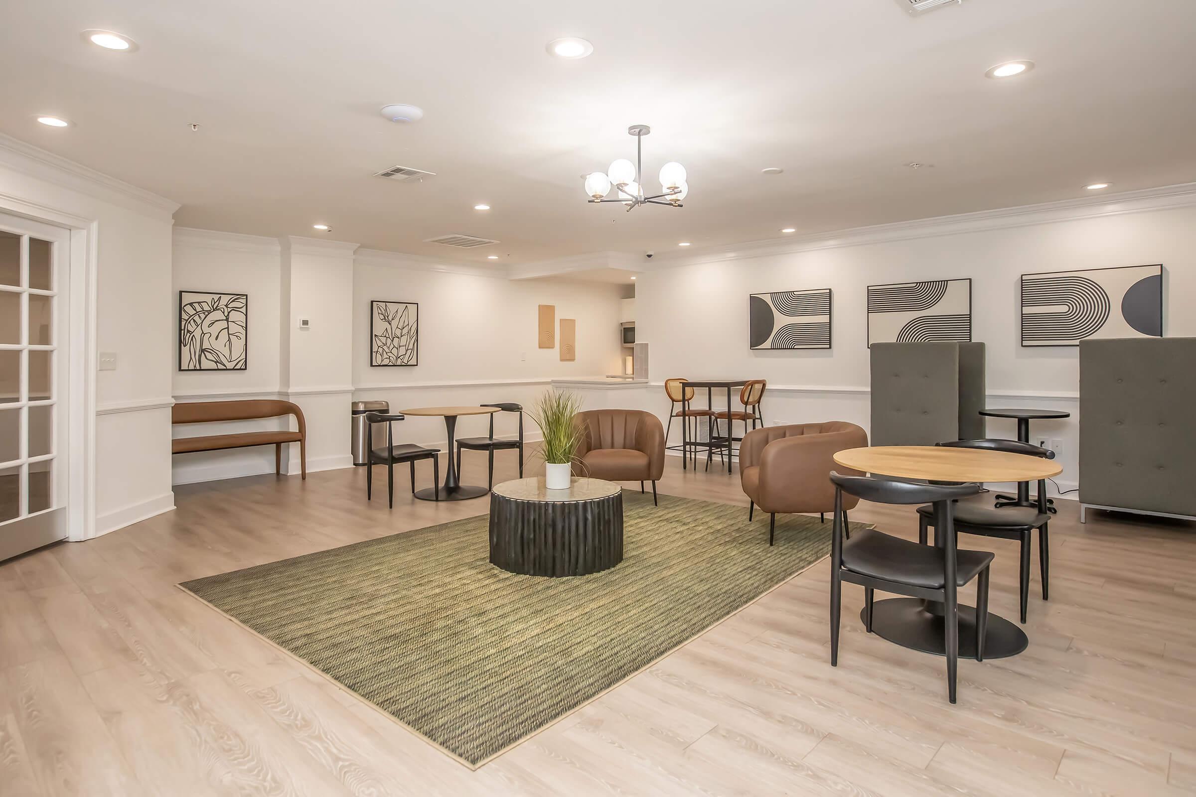 Modern lounge area featuring brown leather seating, round tables, and a green area rug. The walls are adorned with abstract art, and there is a plant centerpiece on the coffee table. The space has bright lighting and a cozy, inviting atmosphere, suitable for socializing or relaxing.