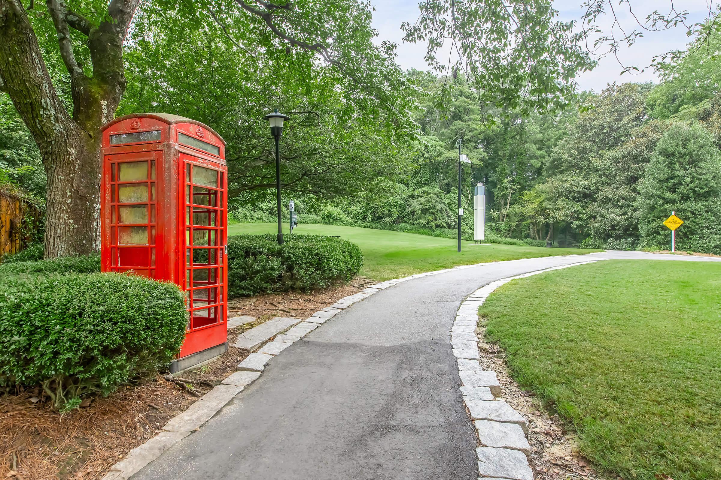A vibrant red telephone box stands beside a winding pathway surrounded by lush greenery. The path is lined with neatly trimmed bushes and leads towards a green park area, with a lamp post visible in the background. Trees provide a serene backdrop, creating a peaceful outdoor setting.