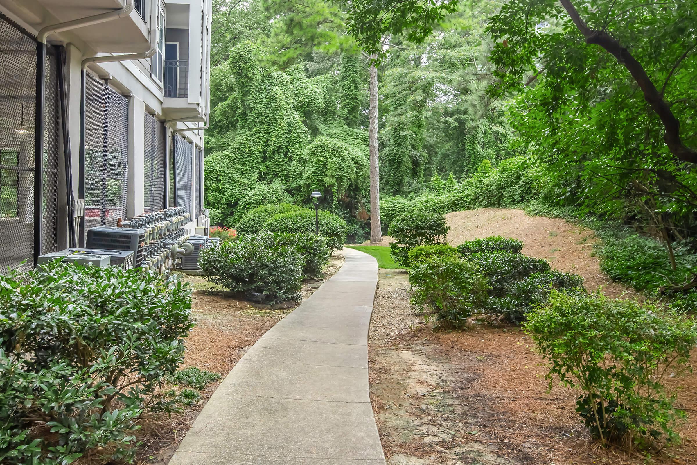 A winding concrete pathway through a landscaped area featuring various bushes and greenery, flanked by a building with air conditioning units. Lush trees and dense foliage create a natural backdrop, contributing to a serene outdoor environment.