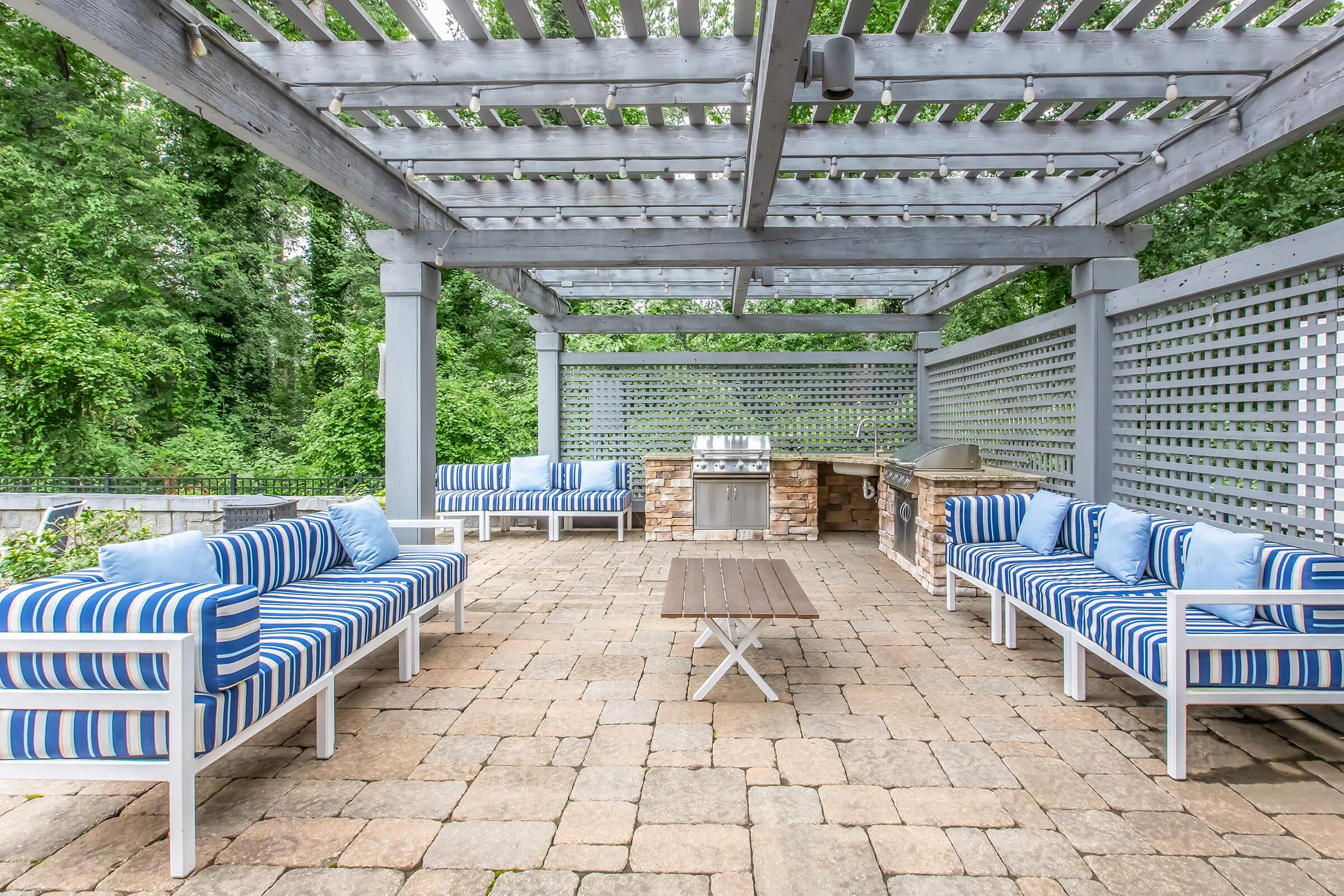 Outdoor patio area featuring striped blue and white seating with cushions, a wooden coffee table, and a stone outdoor kitchen setup. The space is shaded by a pergola and surrounded by green foliage, creating a cozy and inviting atmosphere for relaxation and entertainment.
