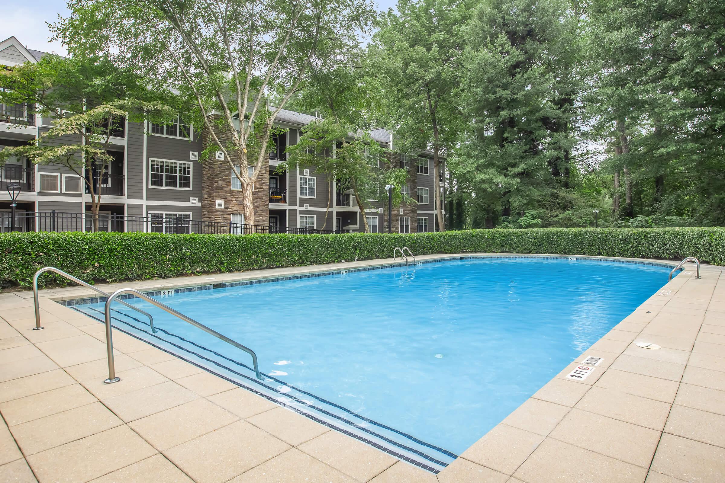 A clear blue swimming pool surrounded by a well-maintained patio and lush greenery. In the background, there are apartment buildings featuring multiple windows and balconies. The scene is bright and inviting, ideal for relaxation and leisure activities.
