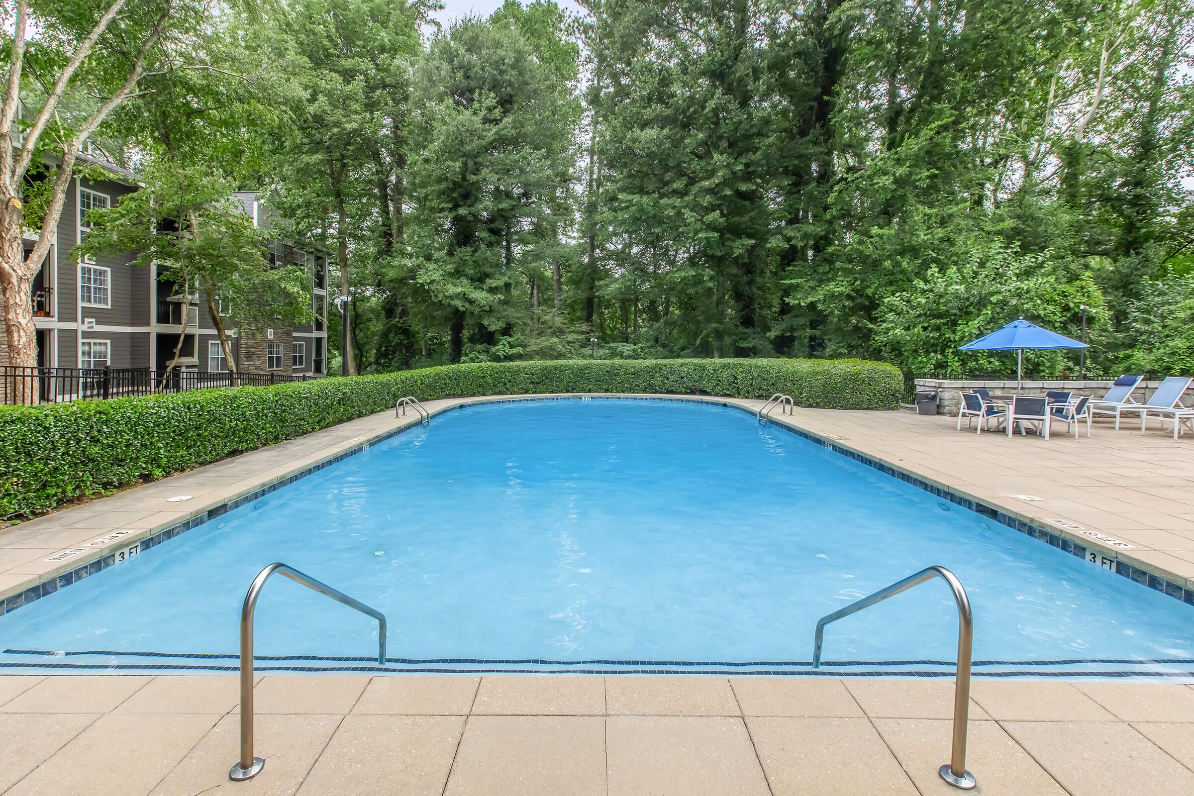 A bright blue swimming pool surrounded by green trees and bushes, with a tan patio area. There are lounge chairs on one side and a shaded umbrella nearby, creating a relaxing outdoor space. The pool's edges have metal handrails for easy access.
