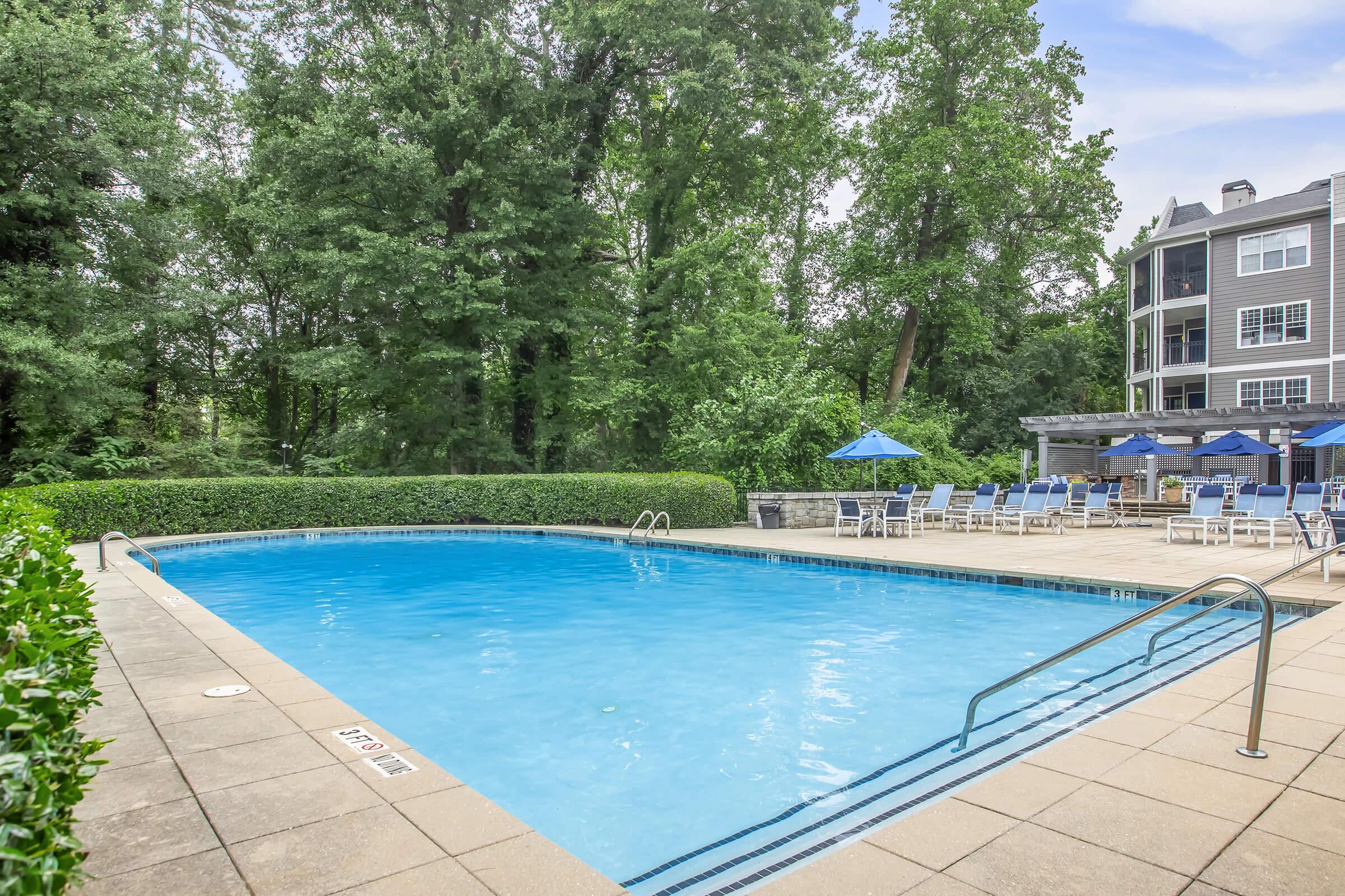 Clear swimming pool surrounded by a stone patio and green hedges. Sun loungers and blue umbrellas are set up nearby, with trees in the background and a multi-story building partially visible. The scene conveys a relaxing outdoor atmosphere ideal for leisure and enjoyment.