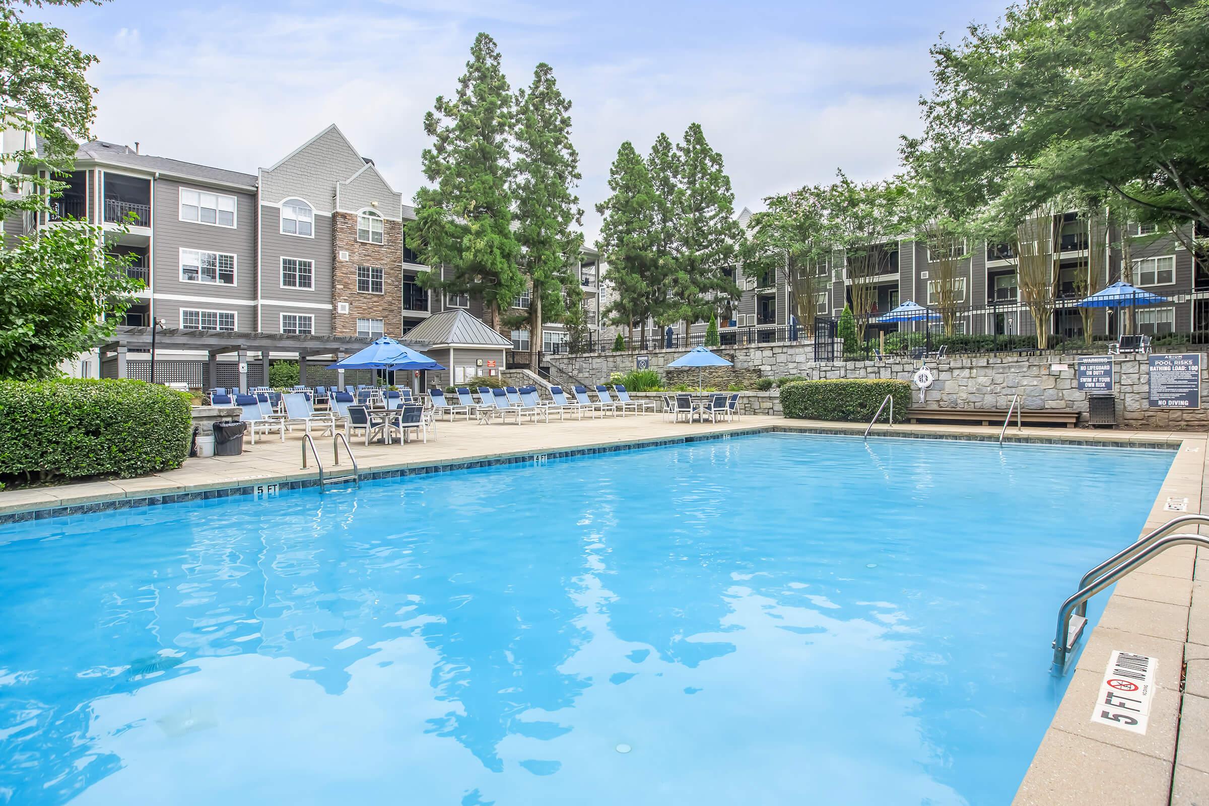 A clear blue swimming pool surrounded by lounge chairs and umbrellas, with lush green trees in the background. Residential buildings are visible on the right side, showcasing a well-maintained outdoor area ideal for relaxation and leisure activities.