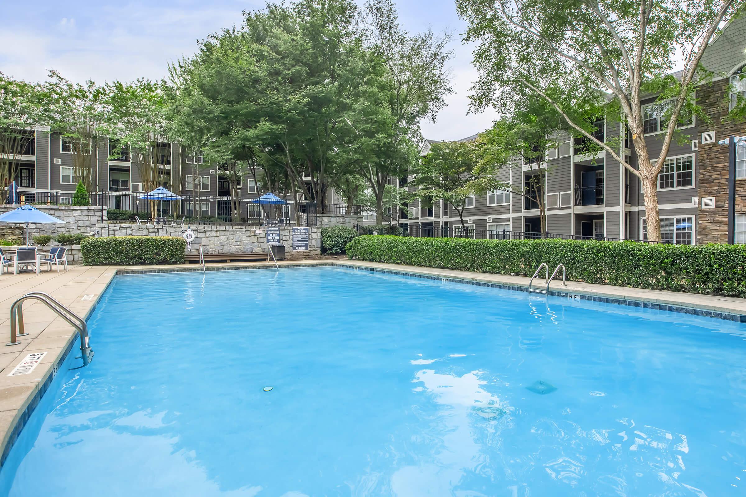 A residential pool area featuring a clear blue swimming pool surrounded by well-maintained green shrubs and trees. There are lounge chairs and blue umbrellas set up for relaxation. In the background, several apartment buildings are visible, creating a serene community atmosphere.