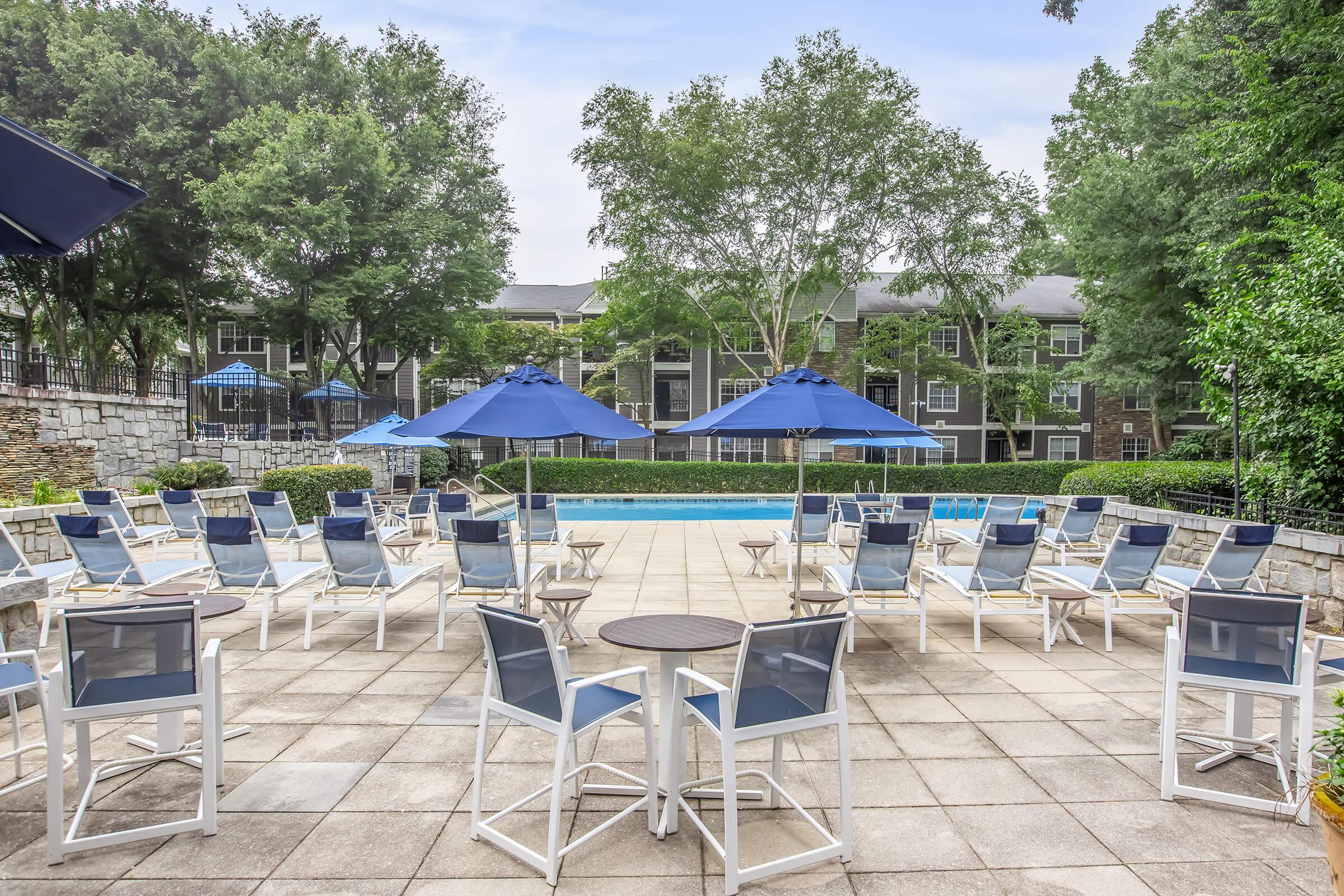 A serene pool area featuring several lounge chairs and round tables, surrounded by green trees and shrubs. Blue umbrellas provide shade, and an apartment complex is visible in the background. The scene conveys a relaxing outdoor atmosphere perfect for leisure.
