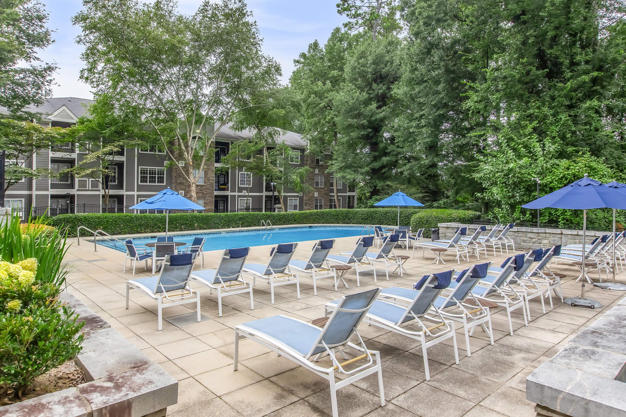 A serene outdoor swimming pool surrounded by lounge chairs and blue umbrellas, set in a landscaped area with lush greenery and trees. The pool area is part of an apartment complex, offering a relaxing space for residents to enjoy.