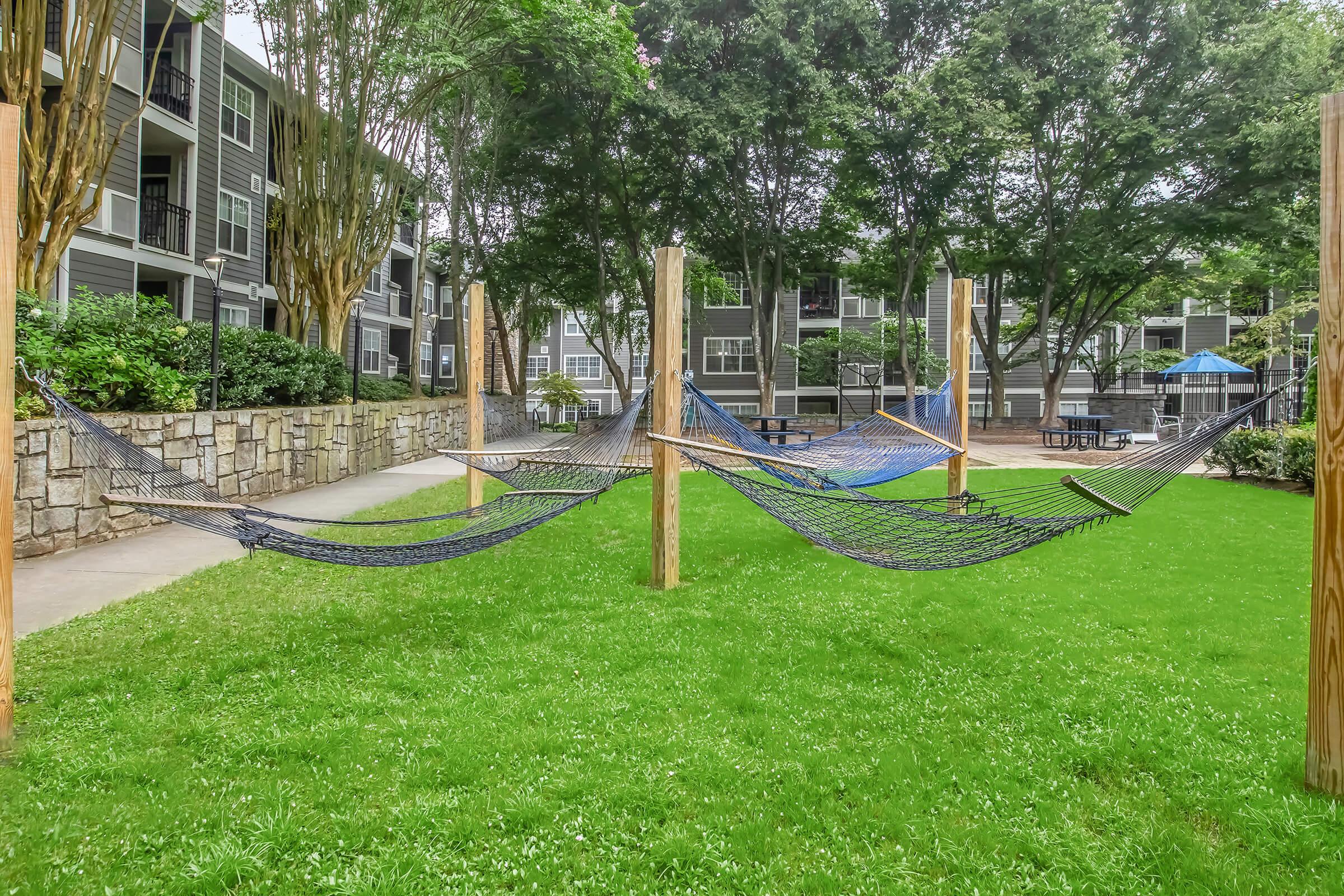 Hammocks arranged in a grassy area surrounded by trees and apartment buildings. The scene features a well-maintained landscape with a walkway, creating a relaxing outdoor space for lounging.