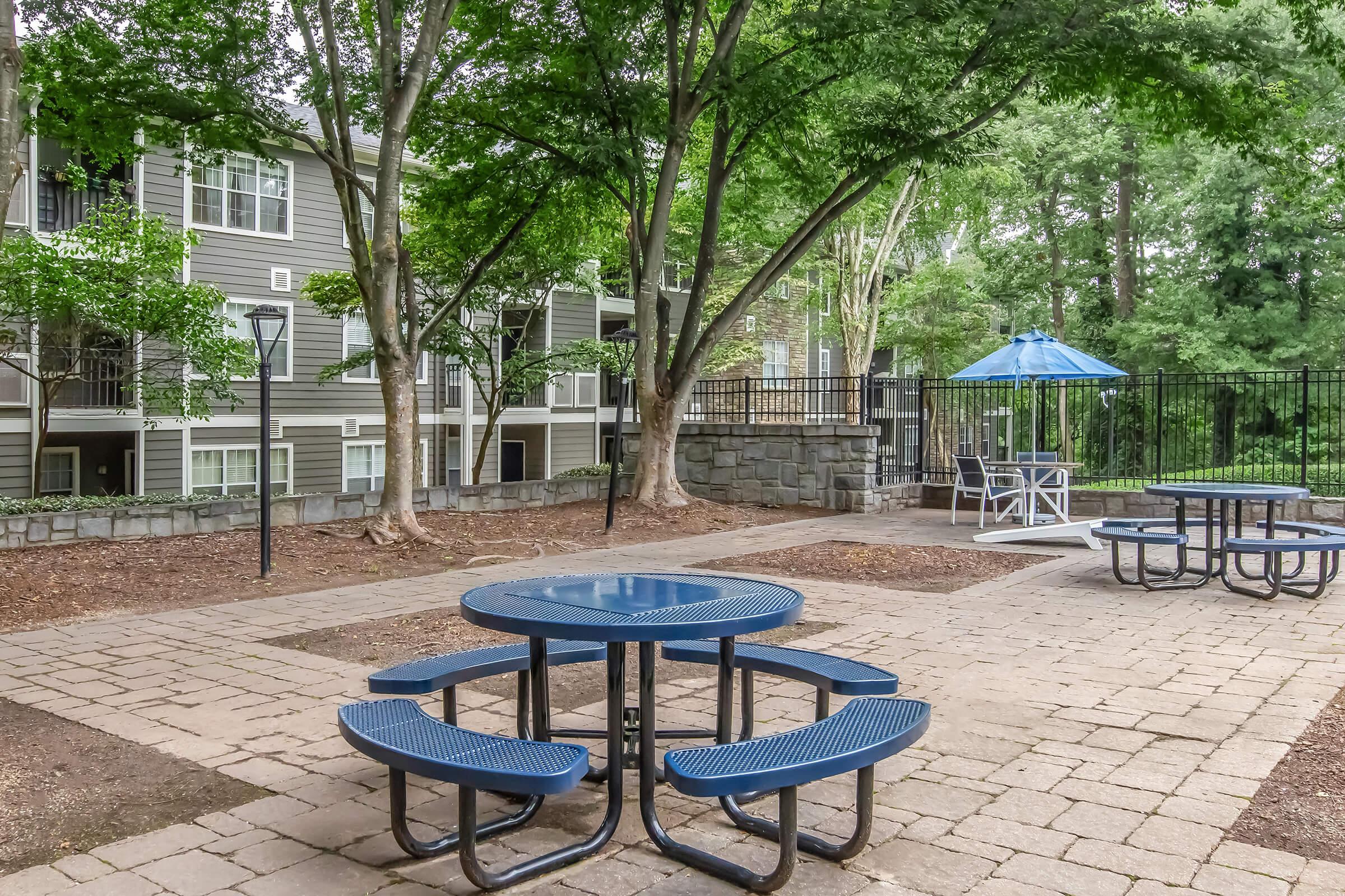 Outdoor communal area featuring blue picnic tables and a seating arrangement under a blue umbrella. Surrounded by green trees and apartment buildings, the space has a paved surface with gravel accents, providing a relaxed atmosphere for residents to gather and enjoy.