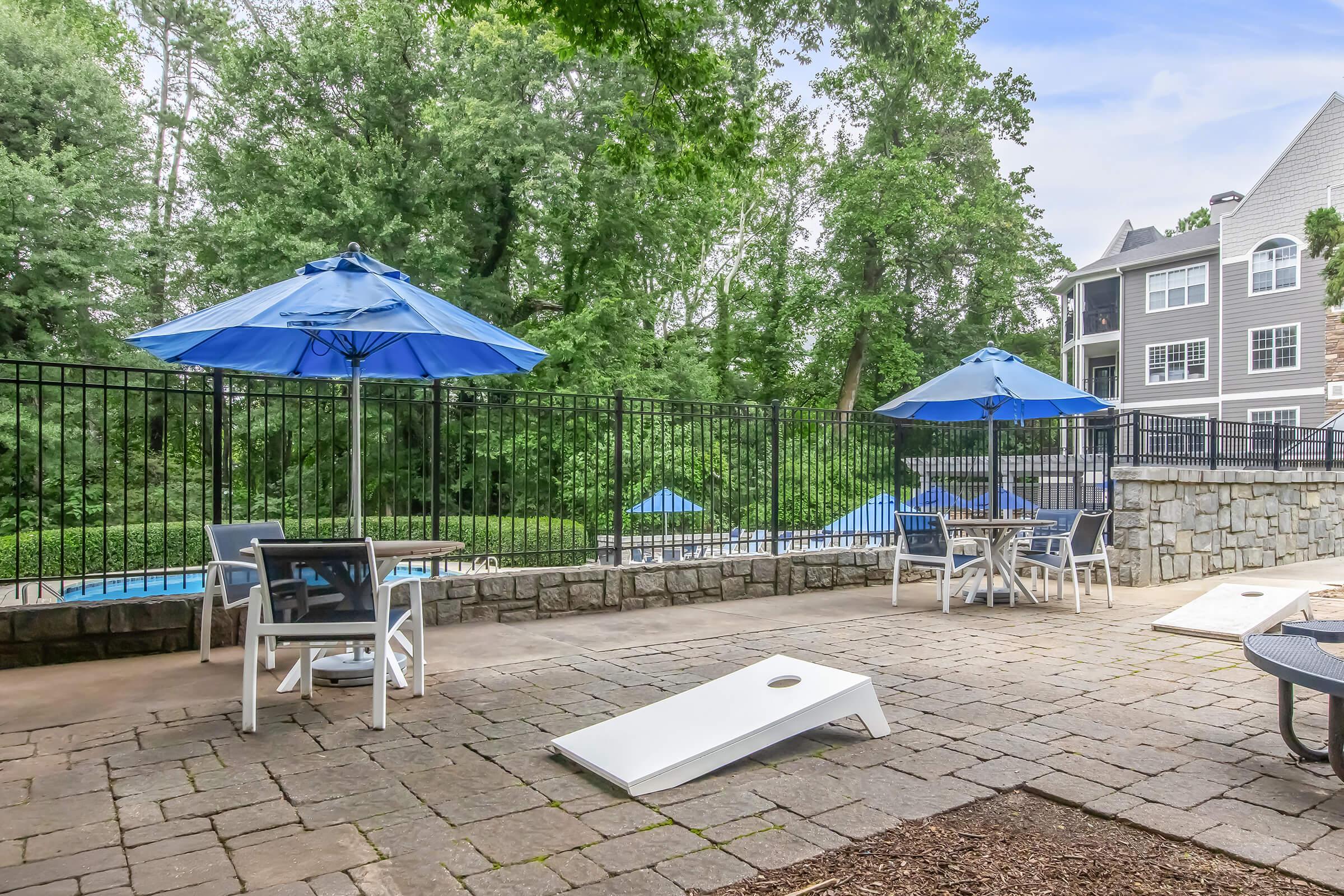A patio area with stone flooring featuring two blue umbrellas over tables, a cornhole game in front, and a view of a swimming pool surrounded by greenery in the background.