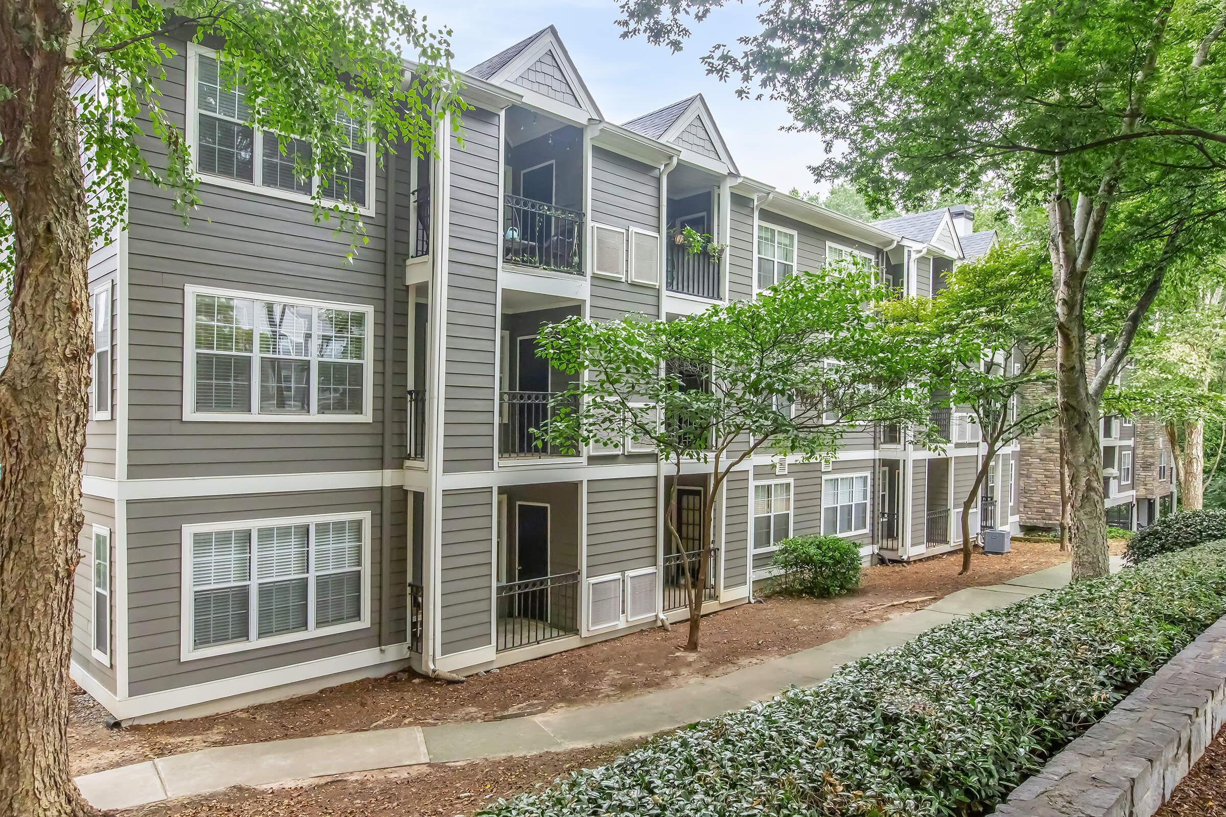 A multi-story apartment building with gray siding and white trim, featuring balconies and large windows. The building is surrounded by greenery, including trees and shrubs. A paved pathway runs alongside the building, providing access to the entrance.