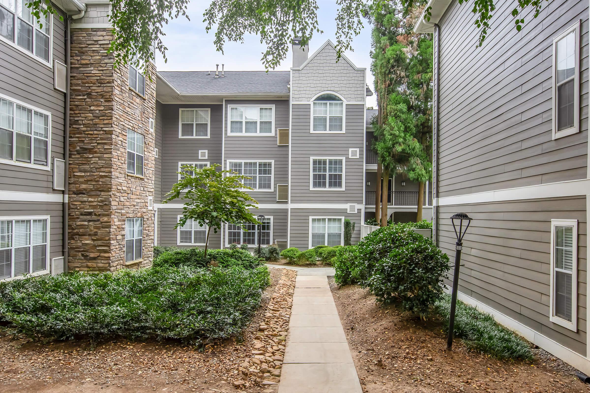 A landscaped walkway leading through an apartment complex, flanked by greenery and trees. The buildings feature a combination of stone and light-colored siding, with multiple windows and balconies visible. The scene is bright and inviting, set against a partly cloudy sky.