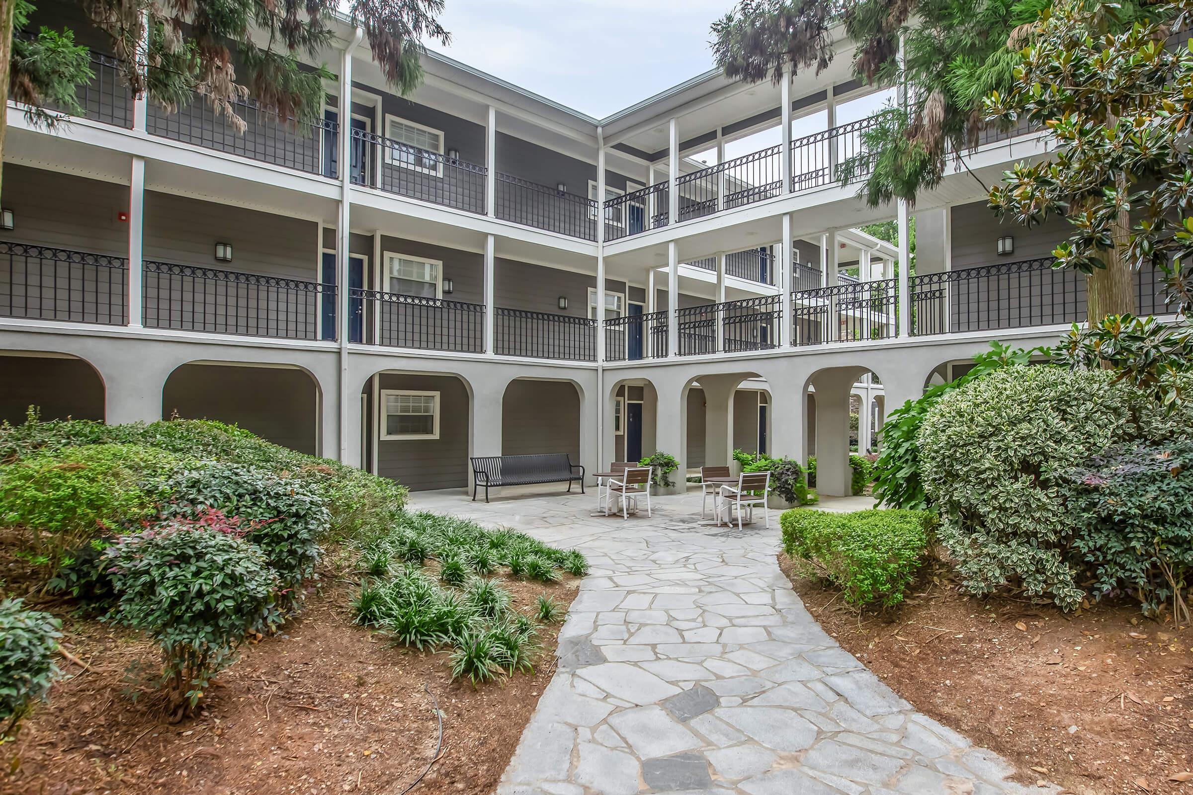 A landscaped courtyard of a multi-story building featuring a stone pathway, surrounded by greenery, with benches and outdoor seating areas visible. The building has multiple entrances with balconies and a light color palette, creating a welcoming atmosphere.