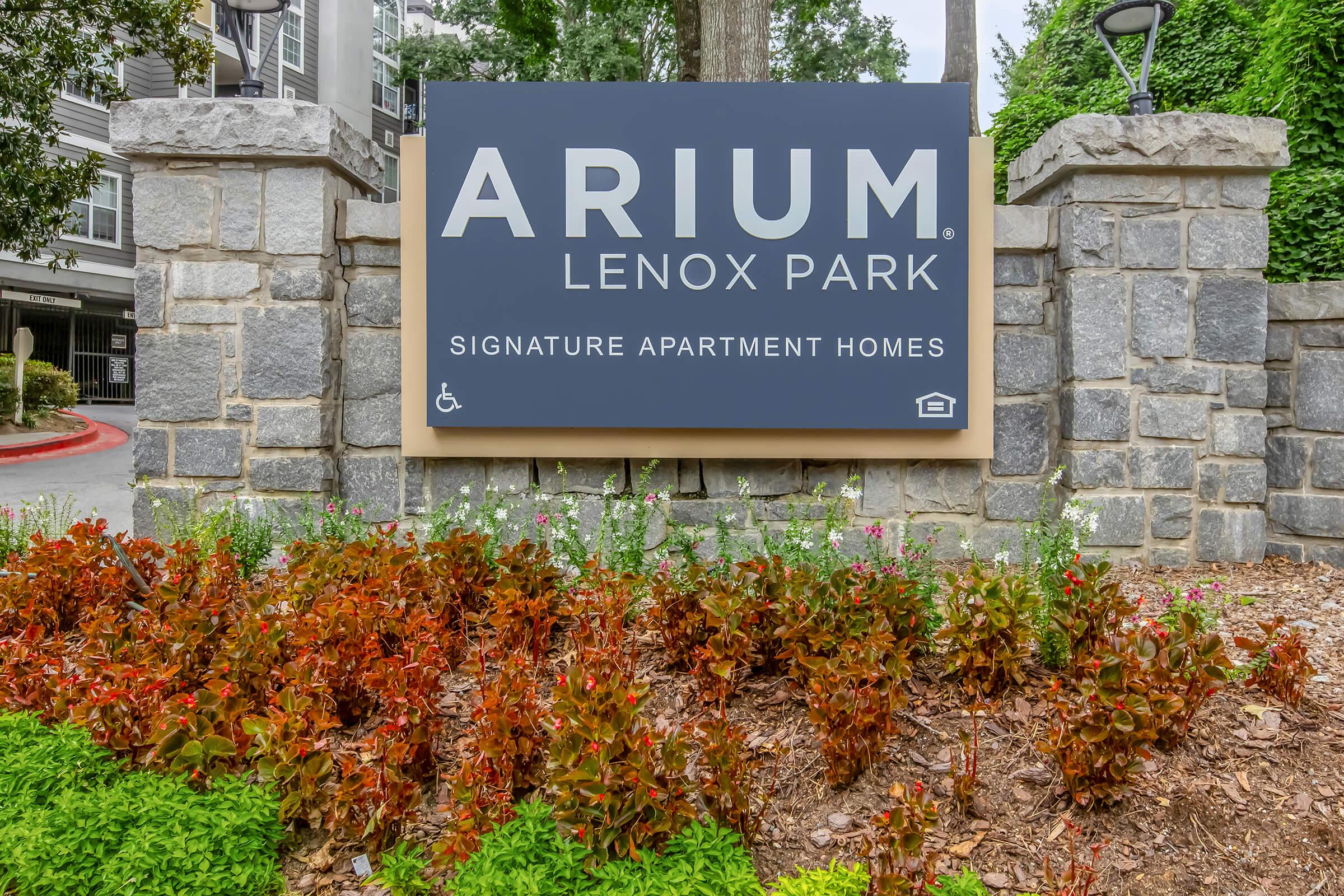 Sign for Arium Lenox Park, featuring the name in bold letters on a dark background with a wooden frame. Surrounded by a landscaped area with colorful flowers and greenery, indicating an apartment complex. The sign also includes a symbol for accessibility.