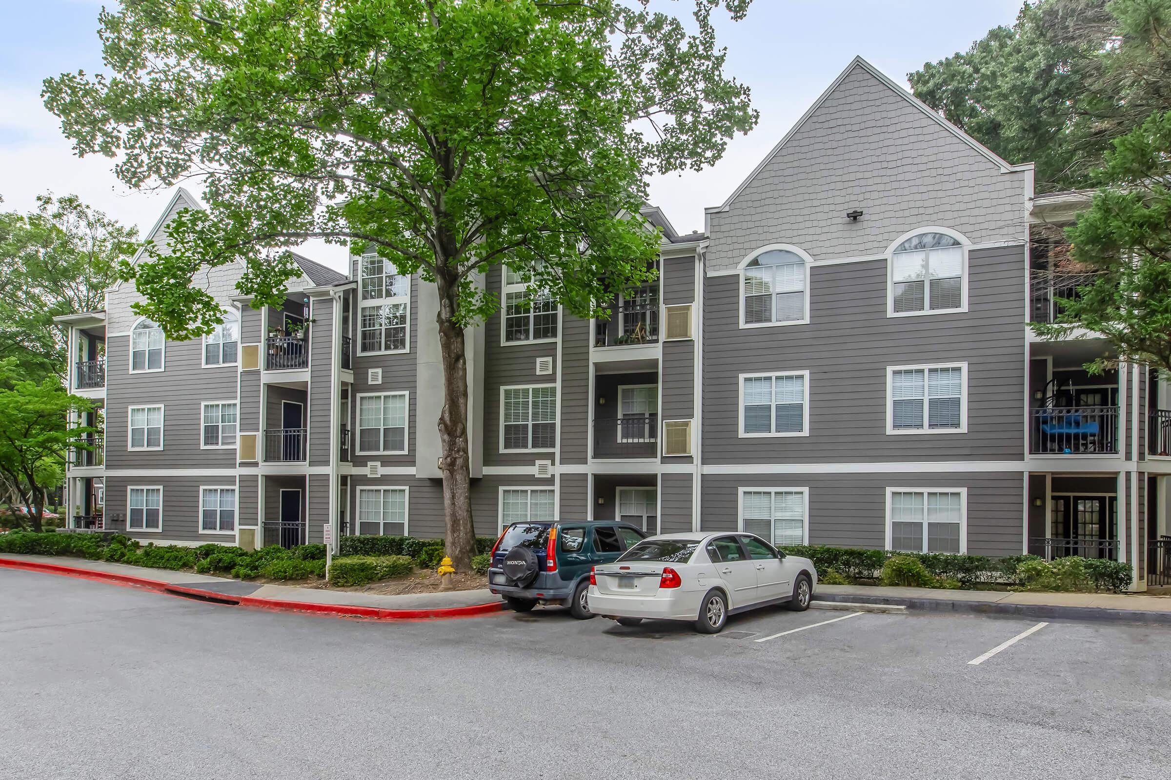 A multi-story apartment building with gray siding and large windows, surrounded by trees and a landscaped area. A parking lot in the foreground features several parked cars, and the sky is partly cloudy.