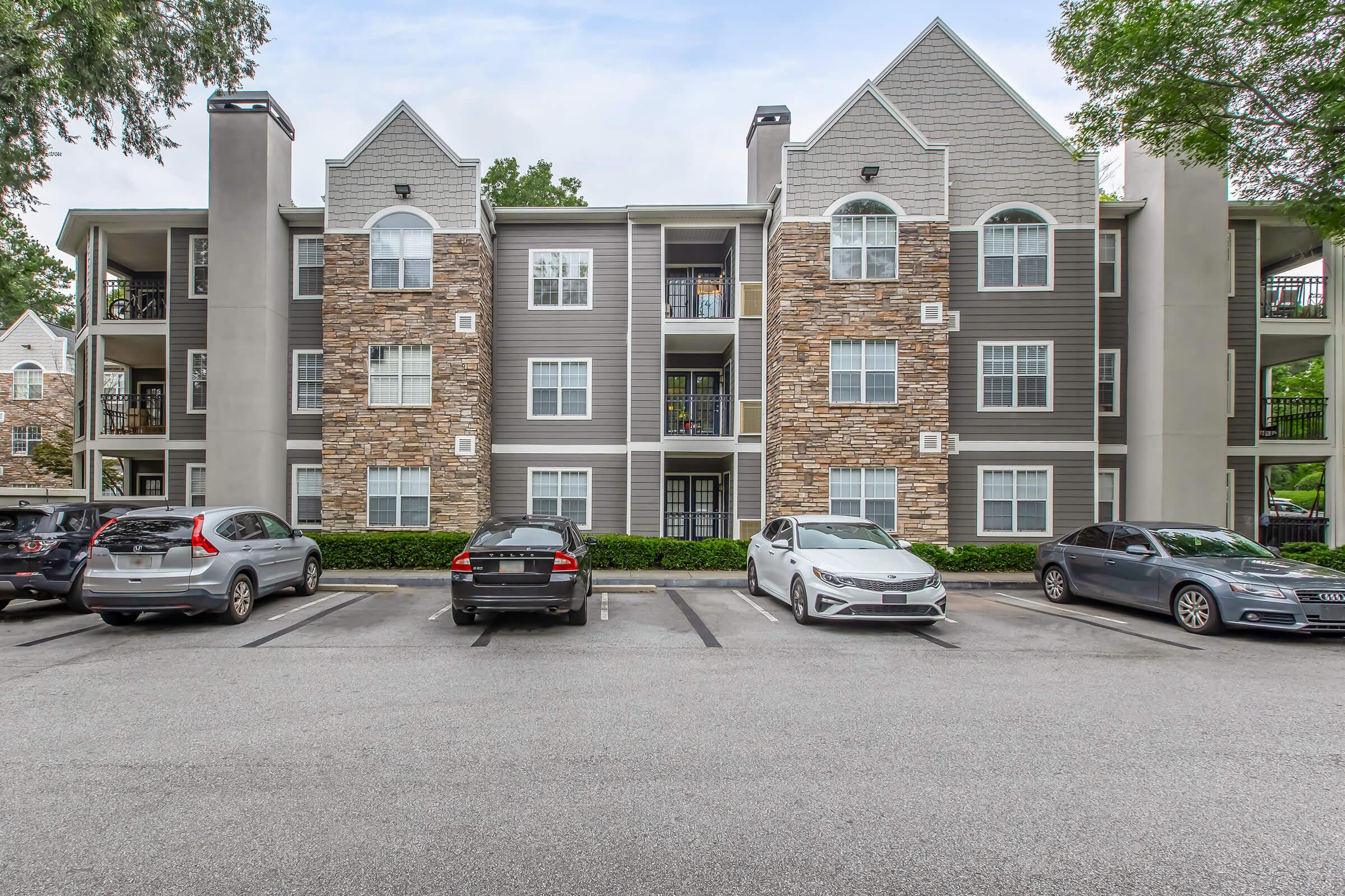 A multi-story residential building featuring a mix of stone and siding exterior, with balconies visible. The parking lot in front has several vehicles parked. Lush greenery surrounds the building, against a backdrop of cloudy skies.