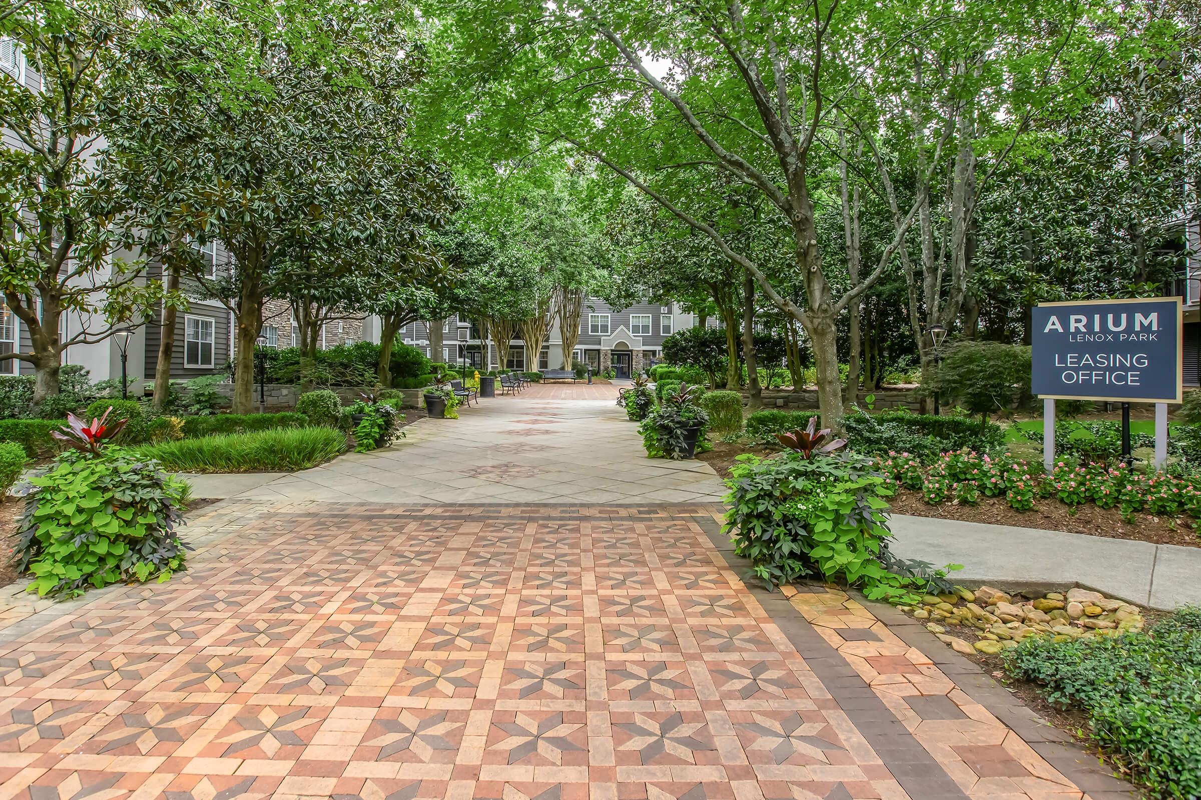 Pathway leading to a leasing office, framed by lush green trees and shrubs. The path features decorative tiles, while a sign for "ARIUM Lenoir Park Leasing Office" is visible on the right. The setting is serene and inviting, surrounded by greenery.