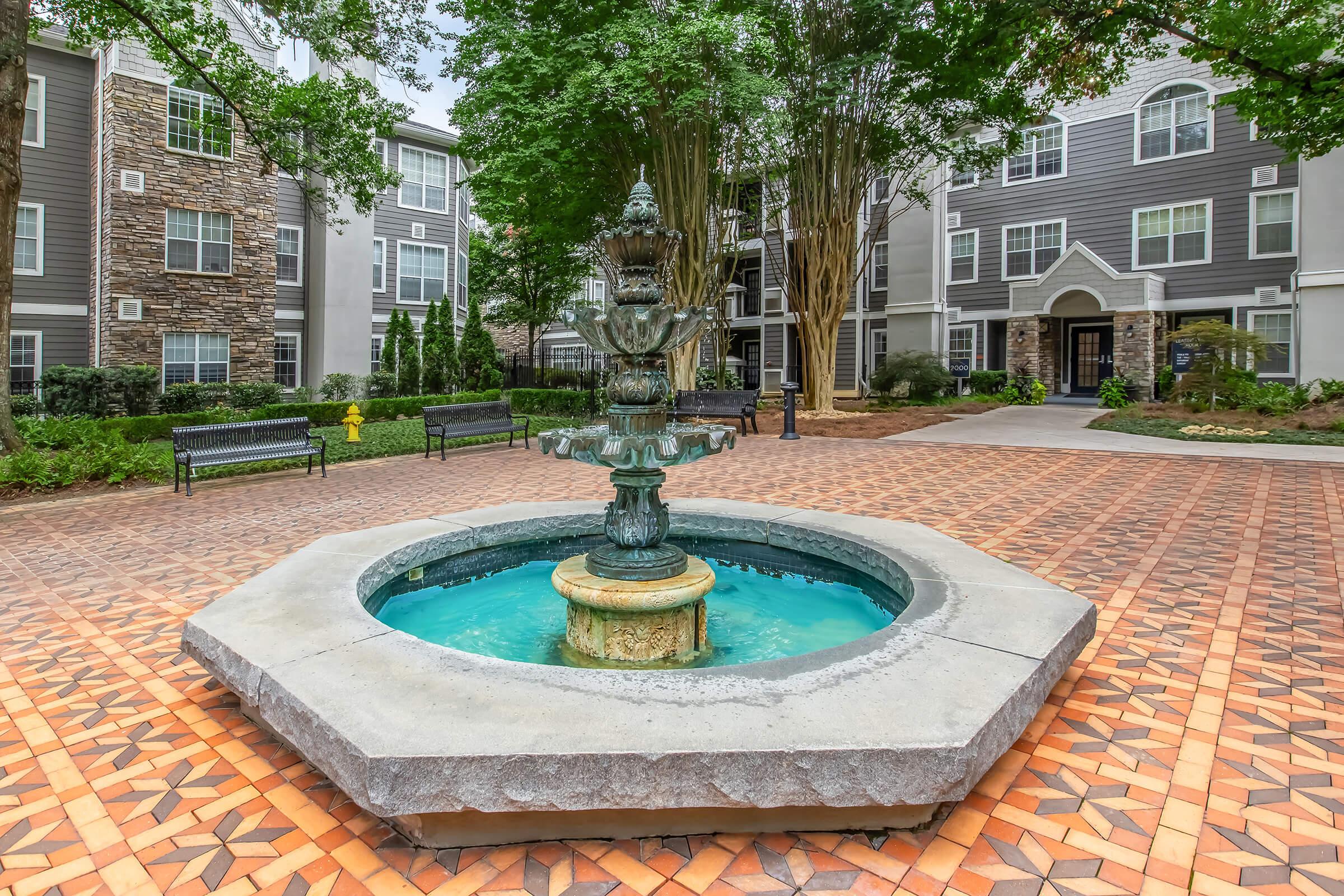 A decorative fountain with multiple tiers sits in a courtyard surrounded by landscaped greenery and modern apartment buildings. The fountain is made of stone and features a circular base with water gently flowing from its top tier, creating a serene atmosphere. Cobblestone pattern surrounds the fountain.