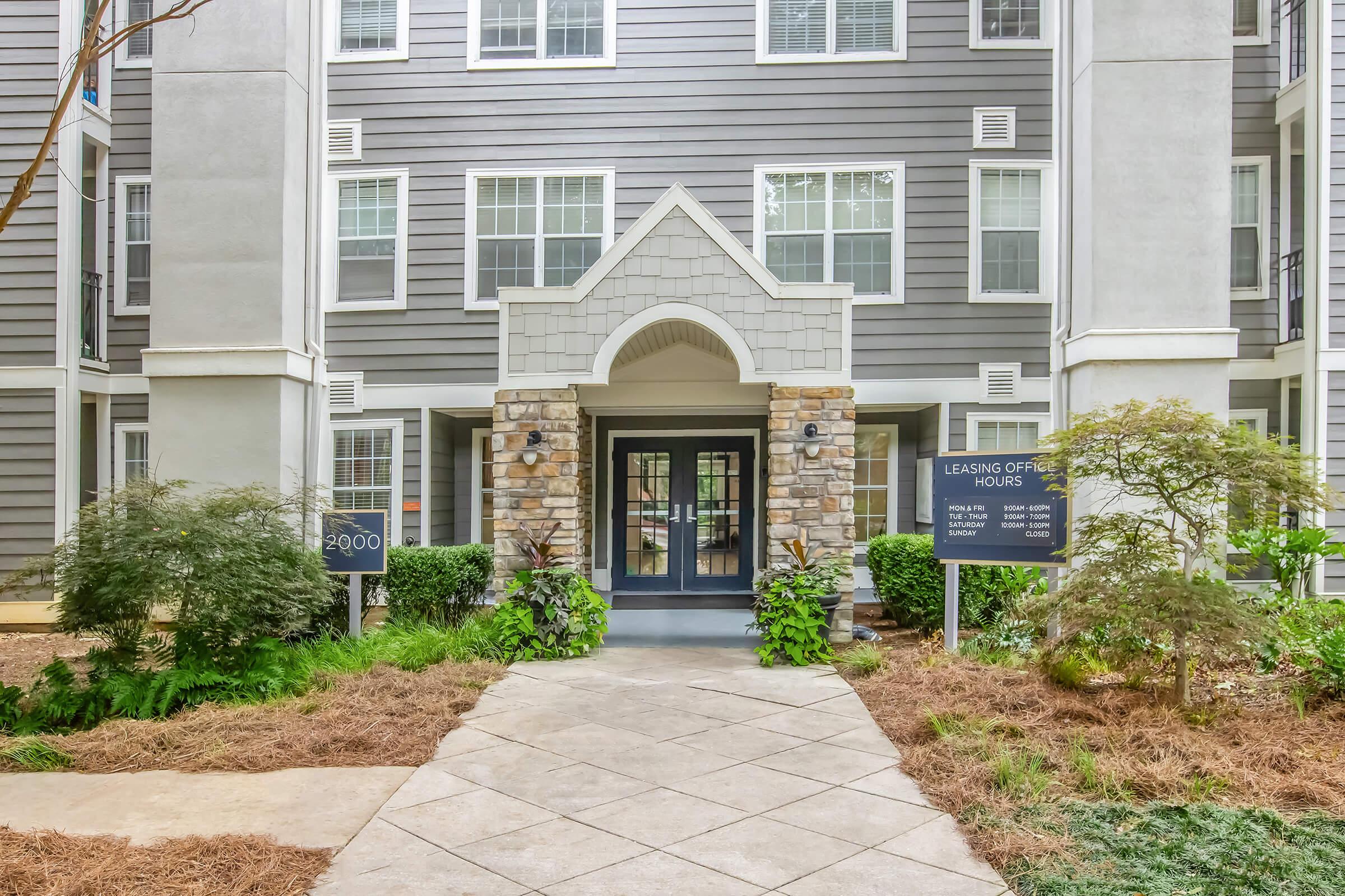 Front entrance of an apartment complex featuring a stone archway, large glass doors, and modern architecture. Flanked by landscaping with shrubs and a sign indicating leasing office hours. The building has multiple stories and a contemporary design with gray siding.