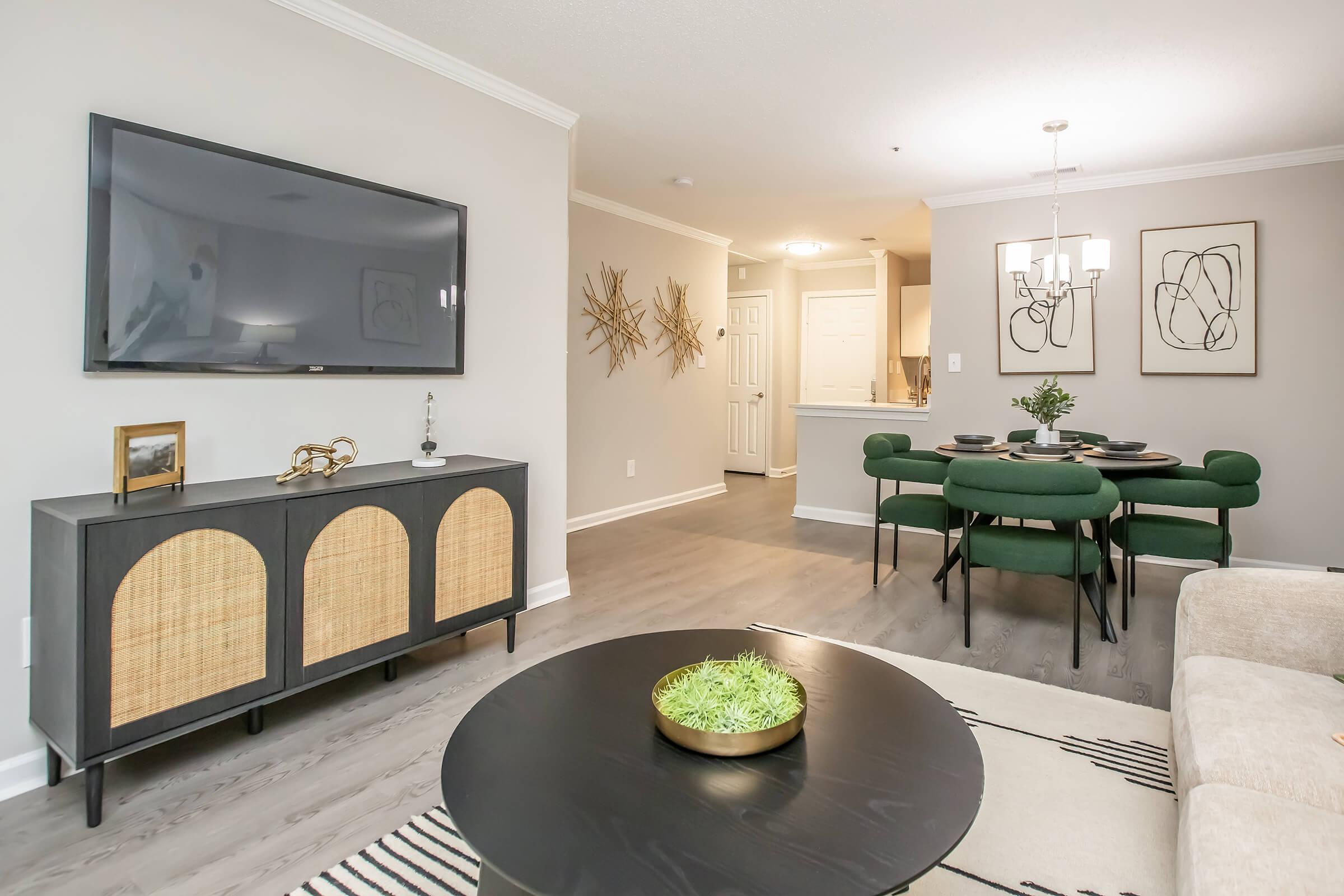 A modern living space featuring a flat-screen TV mounted on a light wall, a stylish black cabinet with wicker accents, a round black coffee table with a decorative plant, and a dining area with a green table and matching chairs. The kitchen is partially visible in the background.