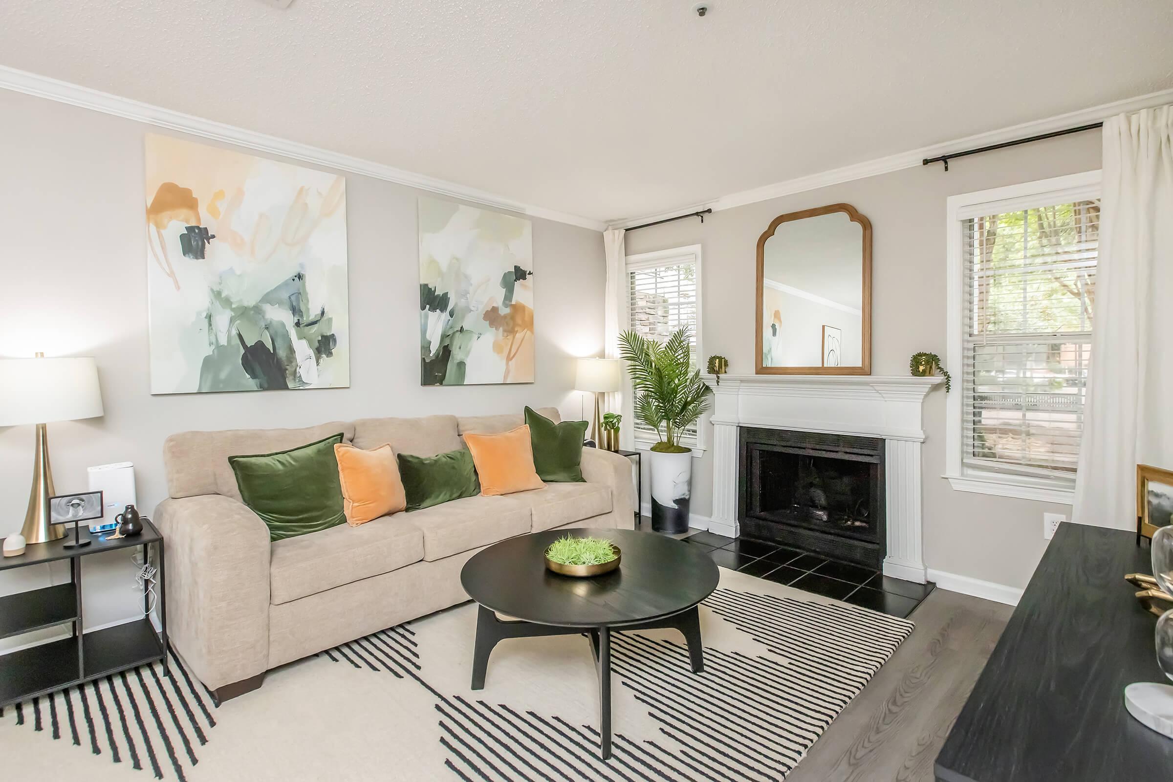 Cozy living room featuring a beige sofa adorned with green and orange pillows, a circular black coffee table, and a decorative plant. The walls are adorned with vibrant abstract artwork, and there's a fireplace with a mirror above it. Large windows allow natural light to fill the space, complemented by a striped rug.