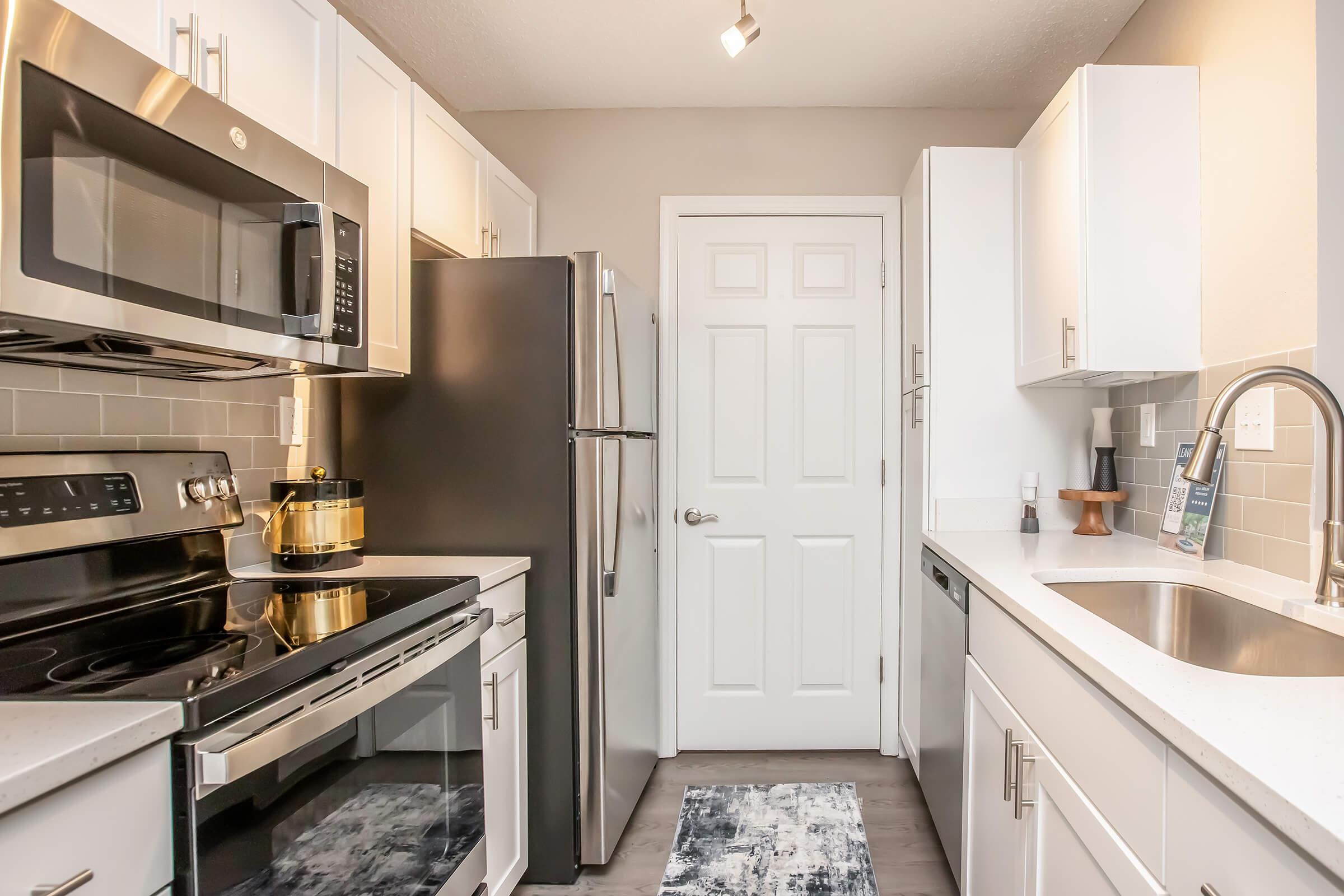A modern kitchen featuring stainless steel appliances including a microwave and oven, a black refrigerator, and a sink. The countertops are light-colored with a subtle texture. White cabinetry provides a clean look, and a gray rug adds warmth to the space. A door is visible in the background.