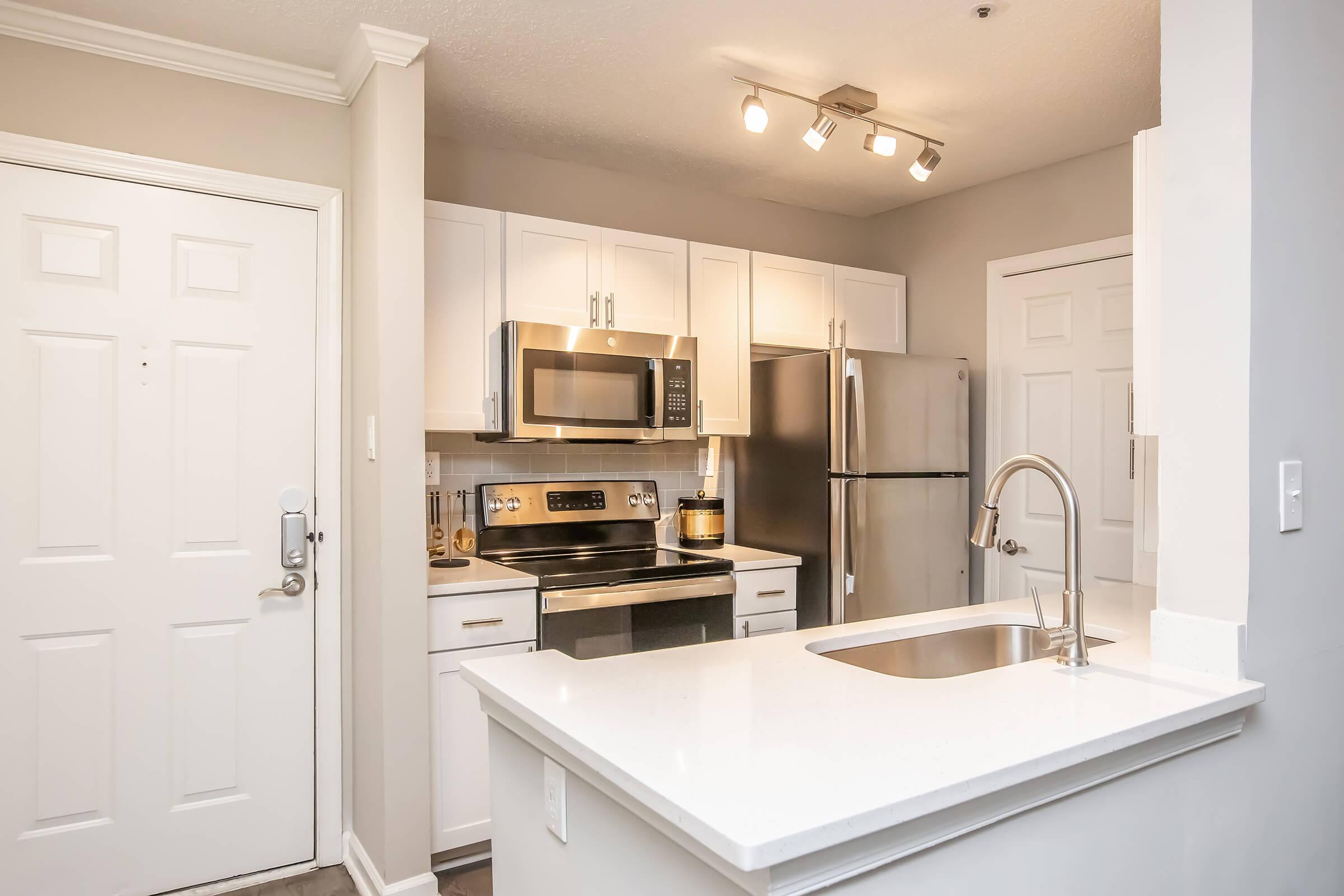 Modern kitchen featuring stainless steel appliances, including a microwave and refrigerator. The white cabinetry complements the light gray walls. A sleek countertop with a sink is positioned in front of the kitchen entrance. Soft lighting highlights the contemporary design.