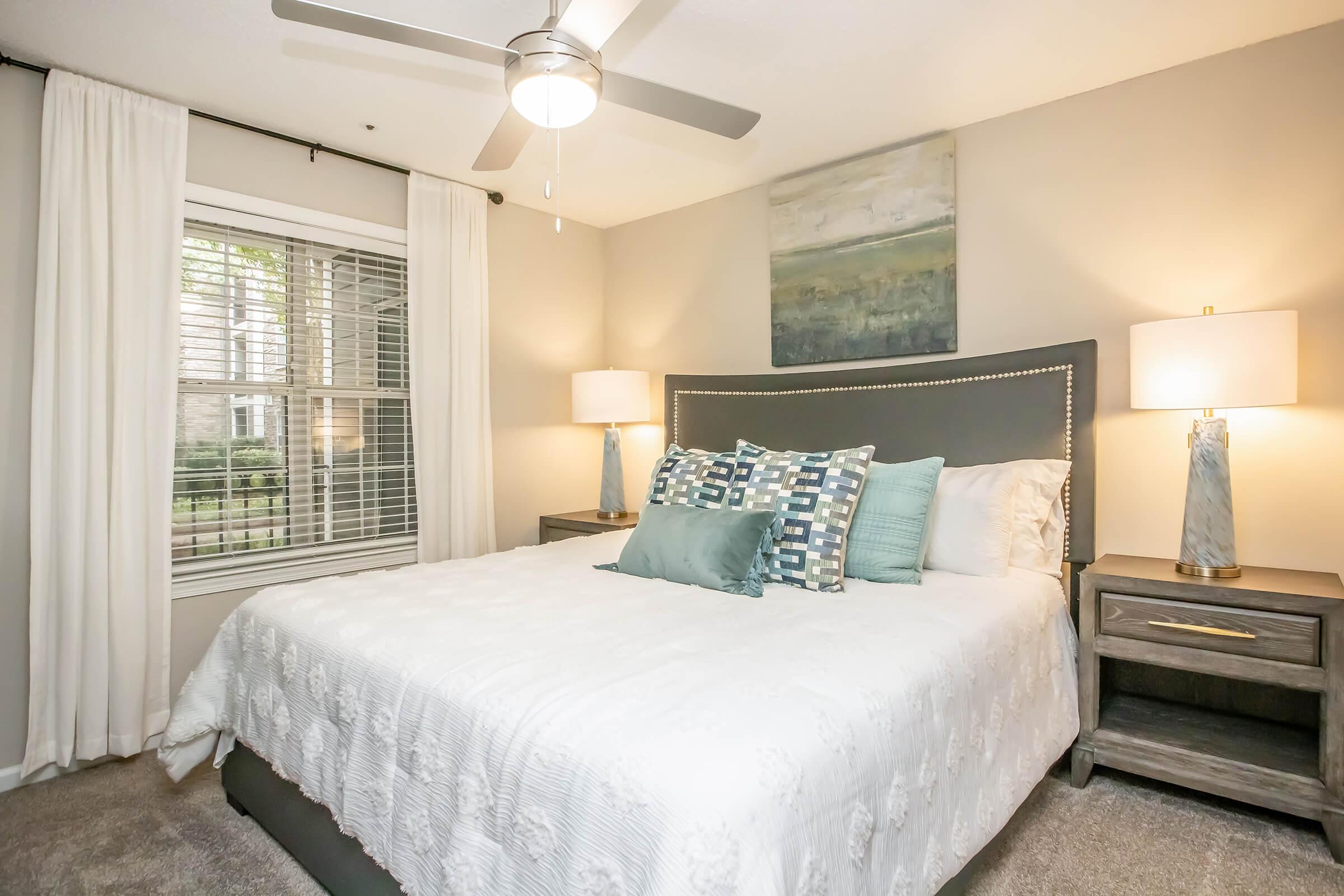 A cozy bedroom featuring a queen-sized bed with a white quilt and decorative pillows. There are two bedside tables with lamps, a ceiling fan overhead, and a large window with sheer curtains allowing natural light. A calming abstract painting is hung above the headboard.