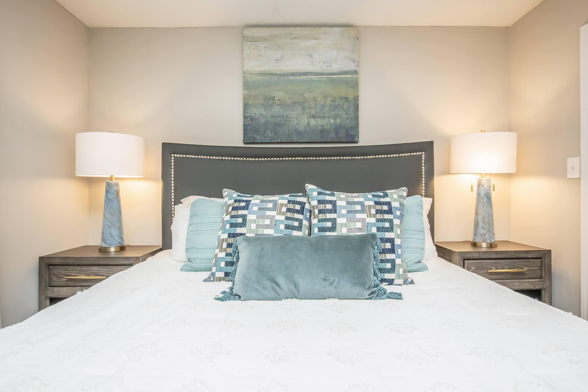 A cozy bedroom featuring a neatly made bed with decorative pillows in shades of blue and gray. On either side of the bed, there are matching nightstands with stylish lamps. Above the bed, there is a serene abstract painting that complements the overall color scheme of the room.