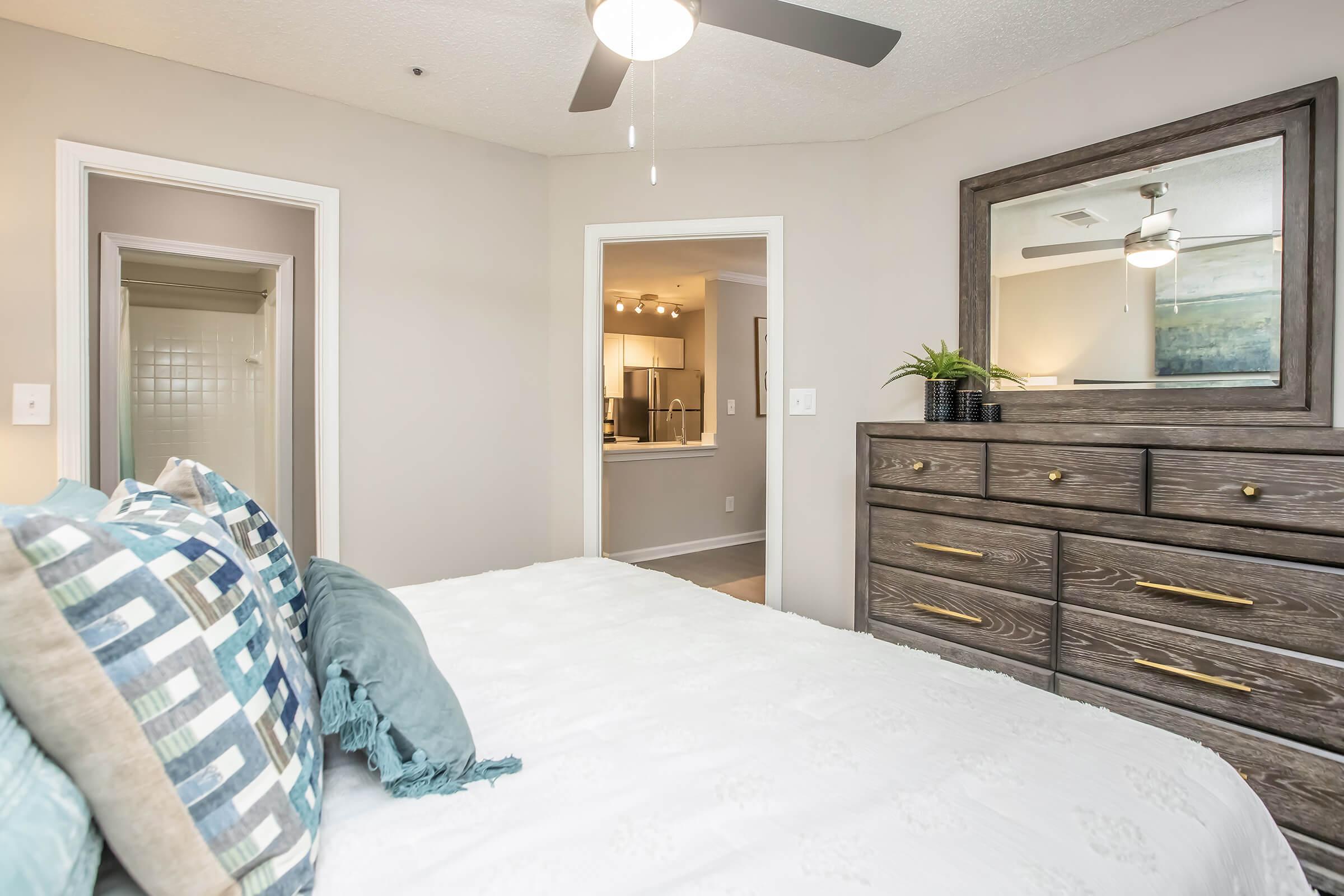 A cozy, modern bedroom featuring a neatly made bed with decorative pillows. A dresser with a mirror and a small plant sits against a wall. Open doorways lead to a bathroom and a living area, while a ceiling fan is mounted above, enhancing the inviting atmosphere.