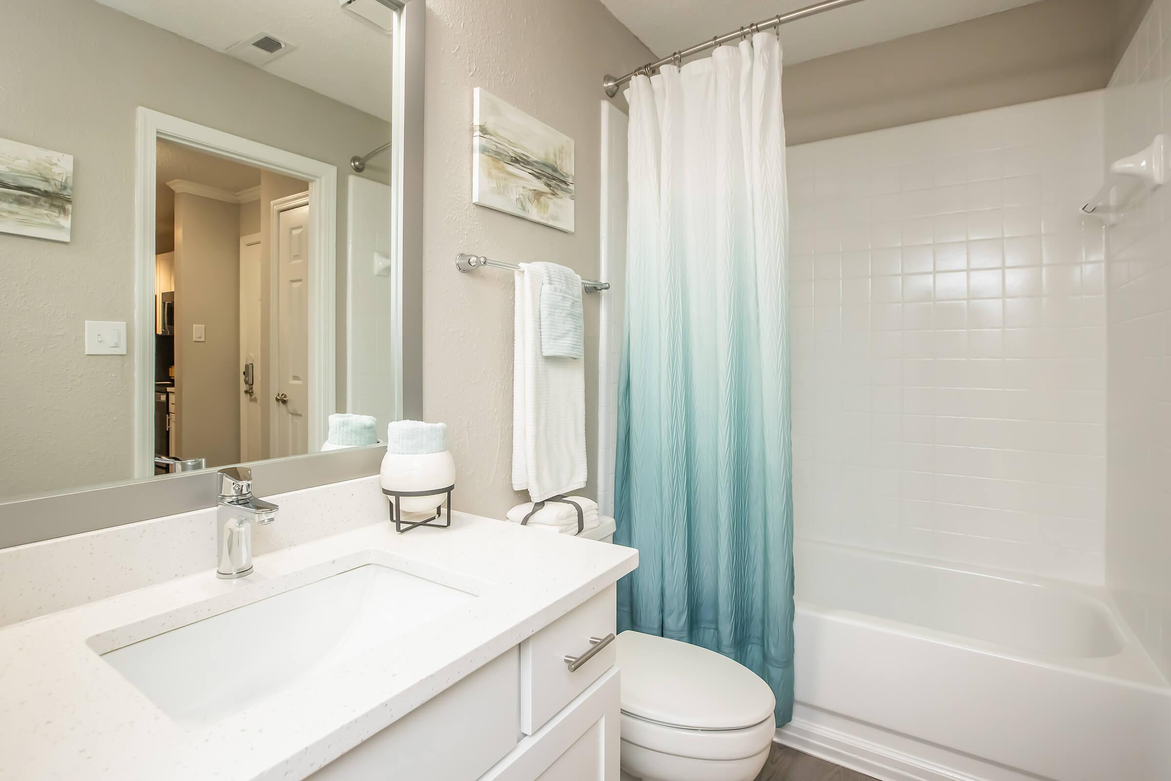 A modern bathroom featuring a white vanity with a sink, a large mirror, and a shower with a blue ombre curtain. The tub is adjacent to the shower, and there are light-colored walls with abstract wall art. A towel rack holds a white towel, and a small decorative item is placed on the vanity.