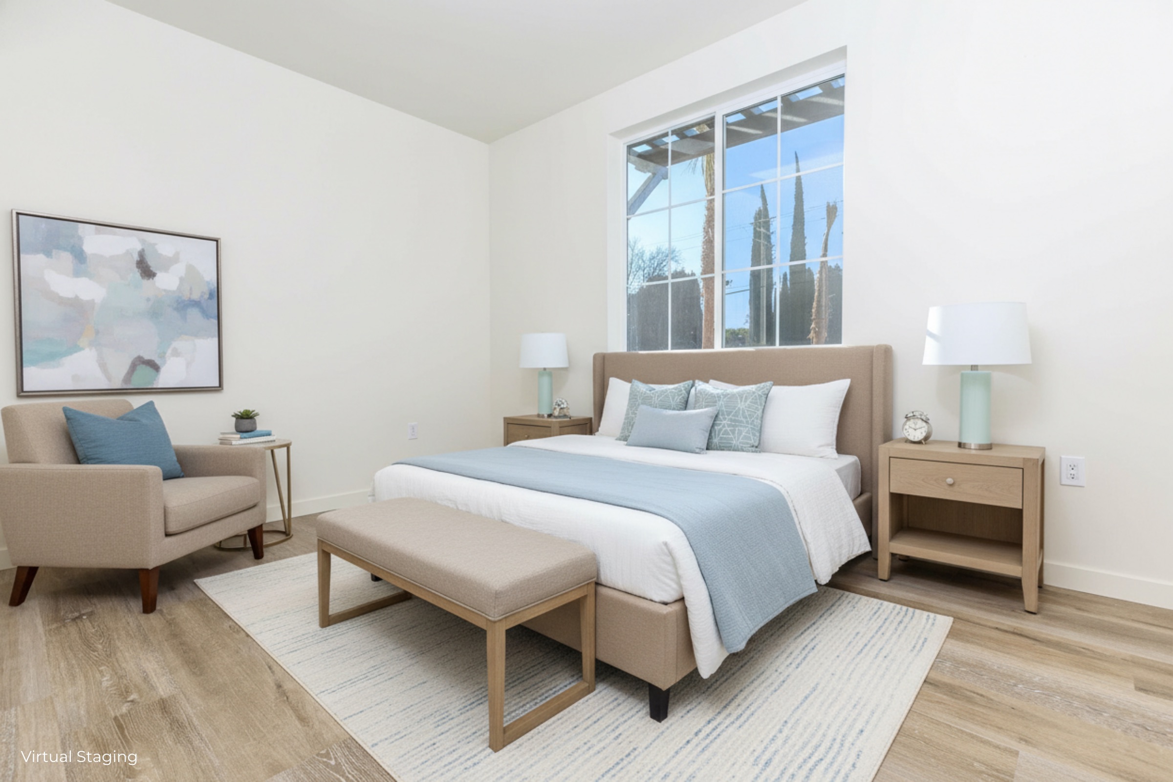 A modern bedroom featuring a beige upholstered bed with light blue accents, a matching bench at the foot of the bed, and wooden bedside tables with lamps. There's a comfortable armchair in the corner and a large window allowing natural light to fill the room, complemented by a light area rug.