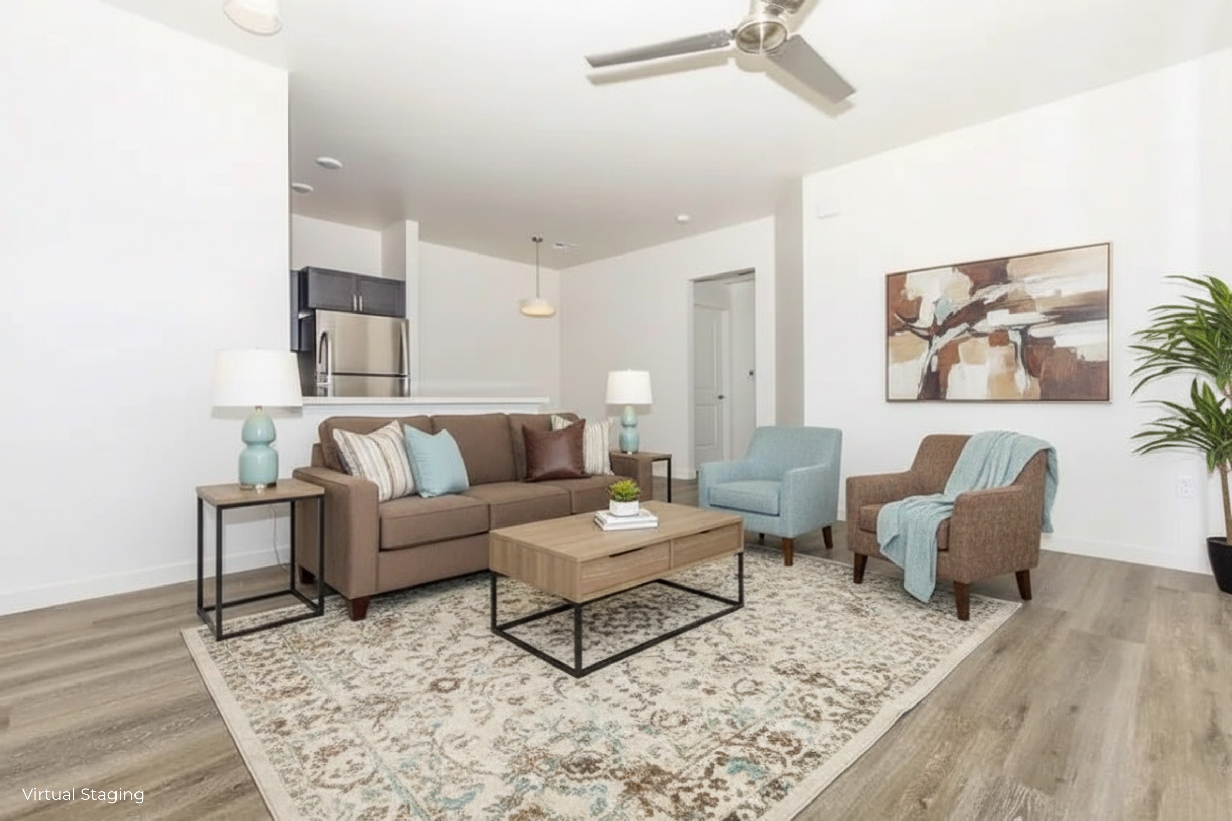 Modern living room featuring a brown couch with decorative pillows, two accent chairs, a coffee table, and a rug. Light-colored walls and wood flooring create a bright ambiance. There are two table lamps on either side of the couch, and a kitchen area is visible in the background.