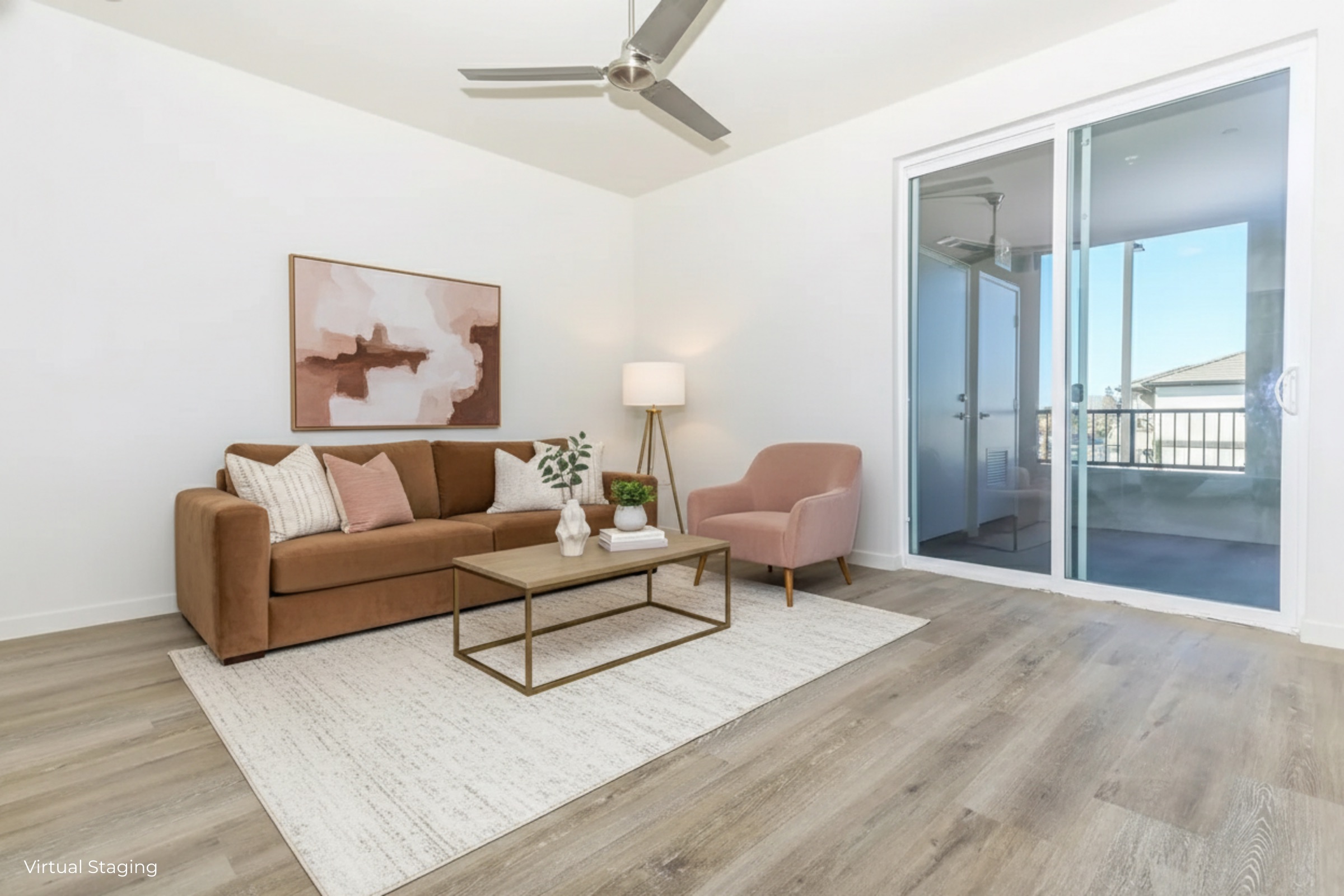 A modern living room featuring a brown sofa with decorative pillows, a pink accent chair, and a coffee table on a light-colored rug. A floor lamp and wall art add a stylish touch. Large glass doors lead to a balcony with natural light streaming into the space.