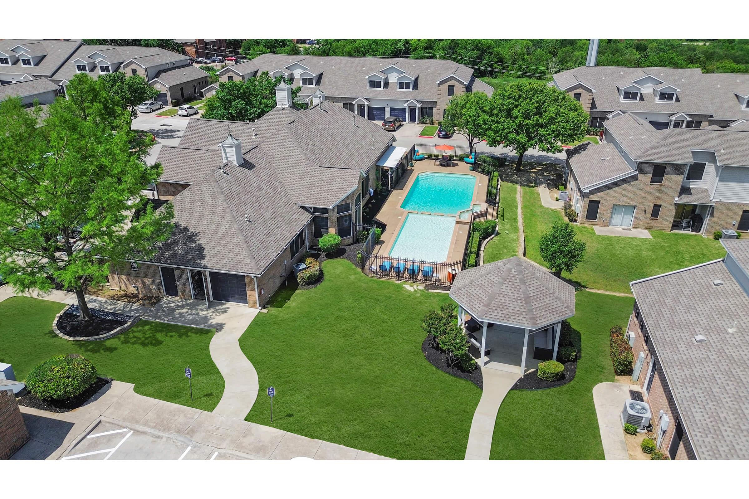 Aerial view of an apartment complex featuring well-maintained buildings, green lawns, a swimming pool surrounded by lounge chairs, and shaded seating areas. Pathways connect the buildings, and there are trees providing greenery throughout the area.