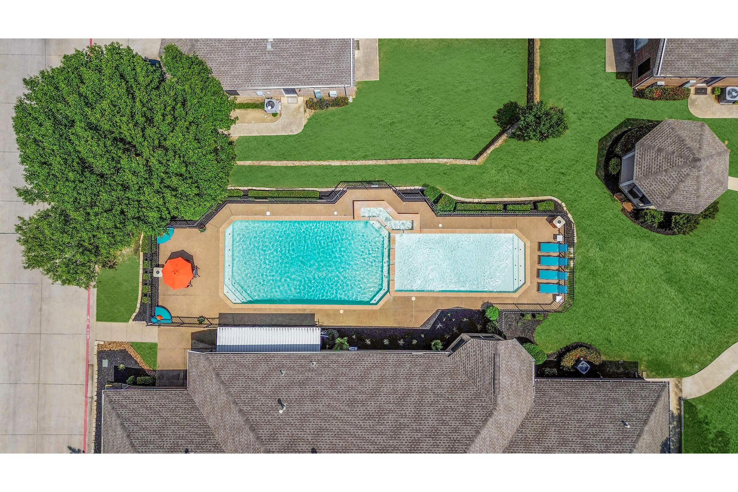 Aerial view of a well-maintained swimming pool area surrounded by green grass. The pool features blue water with an attached hot tub and is bordered by lounge chairs and an umbrella. Nearby, there are landscaped areas and a shaded gazebo, offering a relaxing outdoor environment.