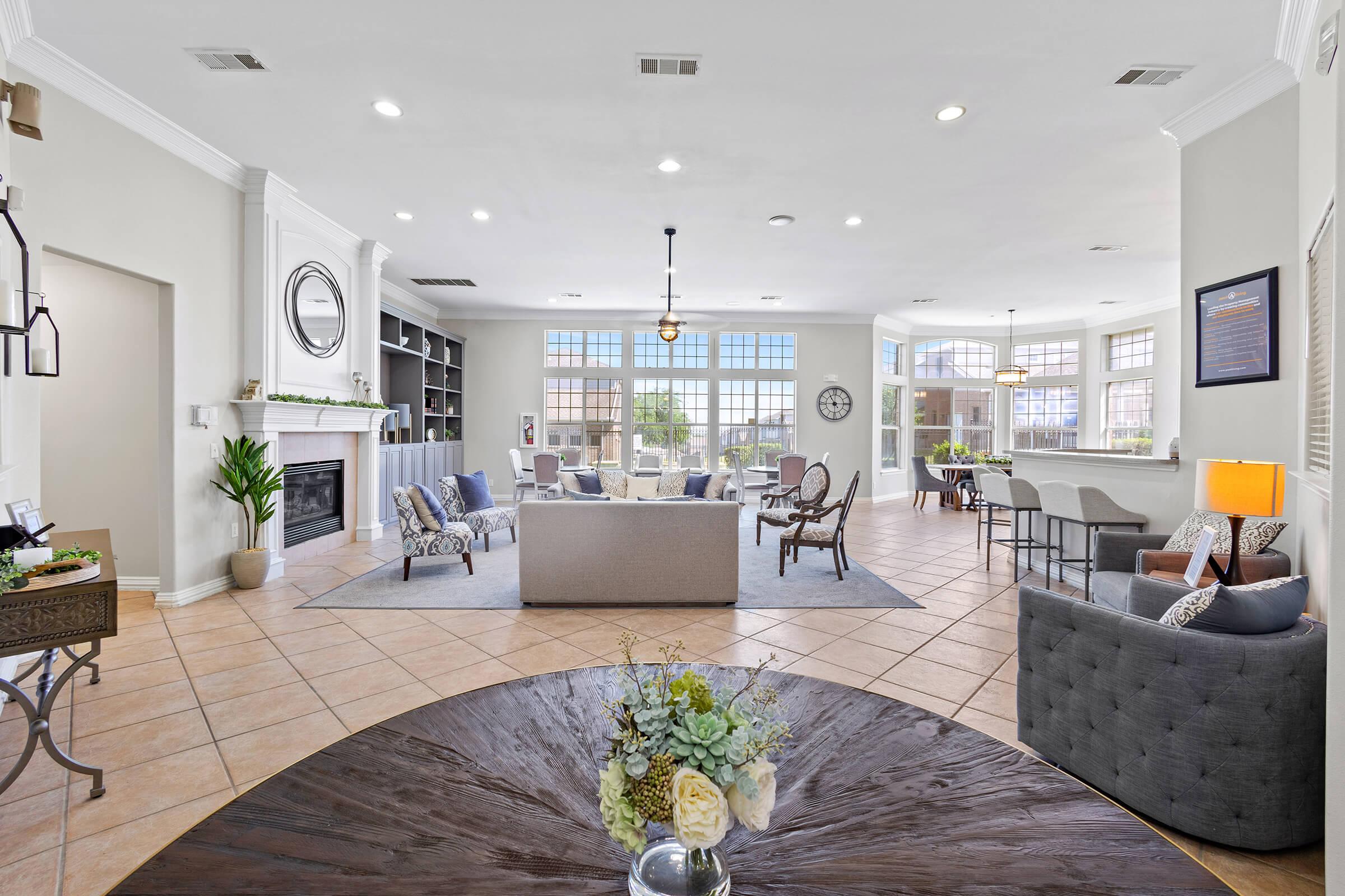 A spacious and well-lit common area featuring modern furniture, including sofas and armchairs, a fireplace, and large windows that let in natural light. Decorative elements like a round coffee table with a floral arrangement add warmth to the inviting atmosphere.