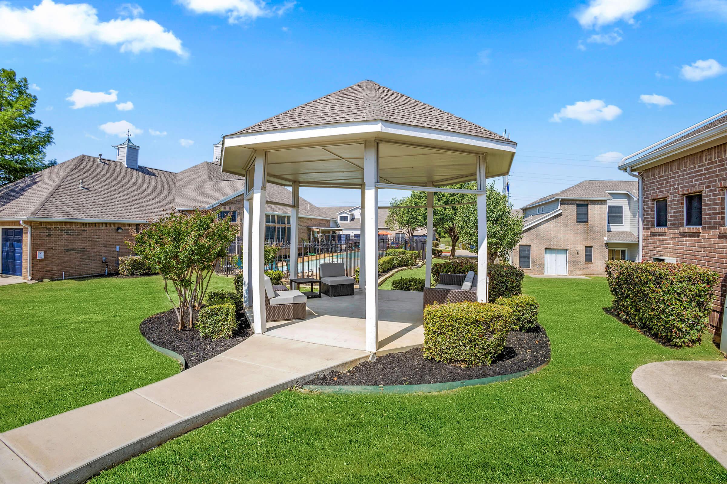 A spacious gazebo with seating is surrounded by well-maintained green grass and shrubs. In the background, there are several residential buildings and a clear blue sky with a few clouds, creating a pleasant outdoor atmosphere.