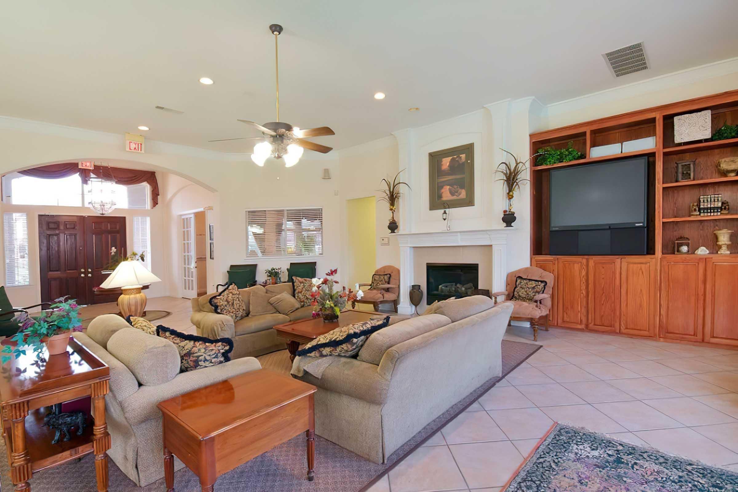 A cozy living room featuring comfortable beige sofas, decorative pillows, and a wooden coffee table. A ceiling fan hangs above, and large windows allow natural light. There is a fireplace with decorative items, a large television on a wooden cabinet, and a welcoming ambiance.