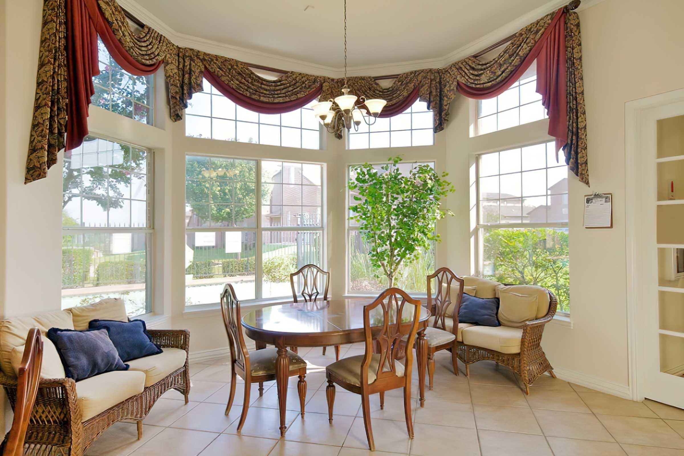 A bright and airy dining area featuring a round wooden table surrounded by elegant chairs. The room has large bay windows adorned with decorative curtains, allowing ample natural light. Potted plants add greenery, and the tiled floor complements the cozy atmosphere.
