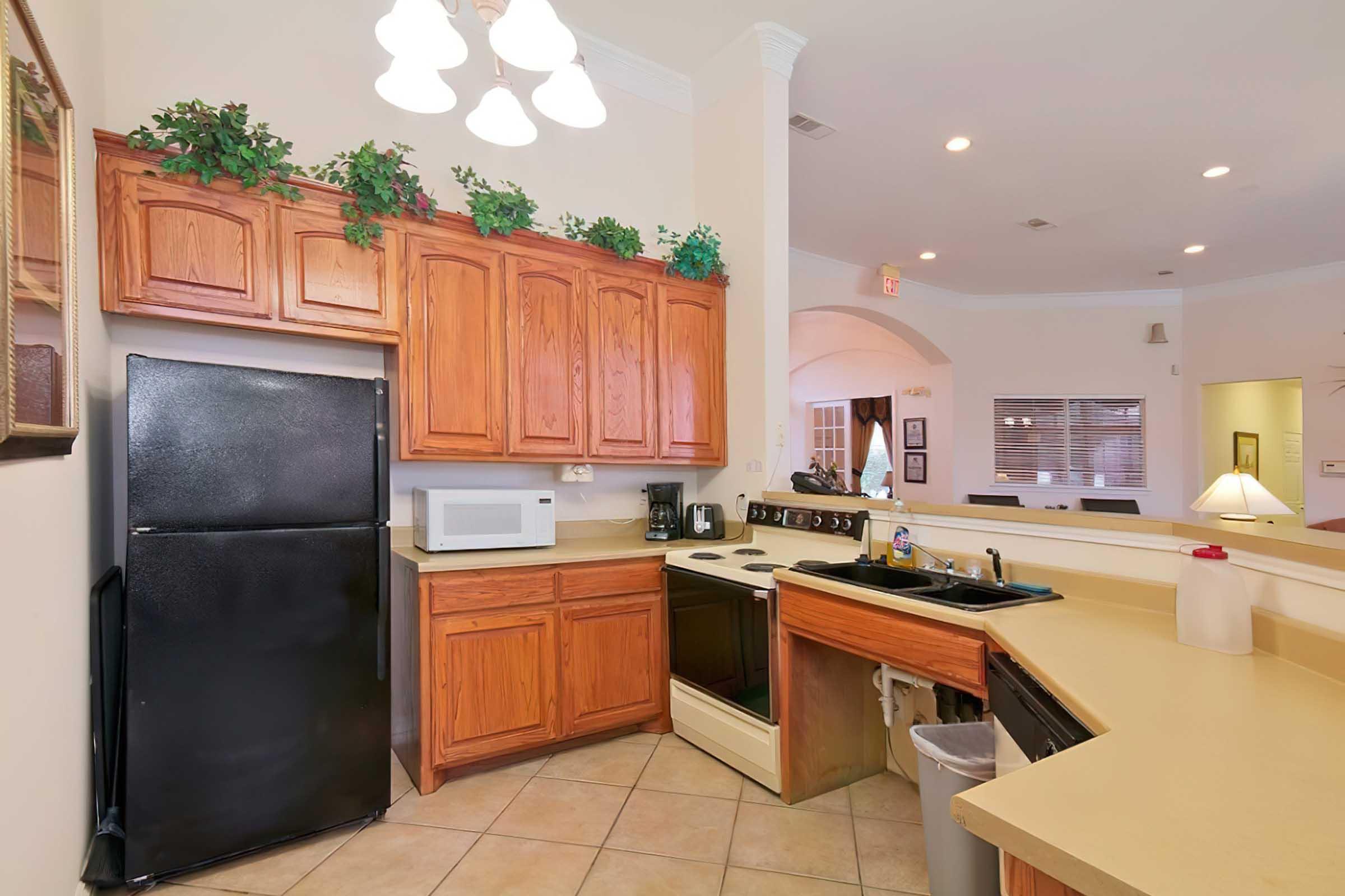 A modern kitchen featuring wooden cabinets, a black refrigerator, a microwave, and a black coffee maker. The countertop is beige with a sink and a trash can, while potted plants adorn the cabinets. The room has warm lighting from pendant lamps, creating a welcoming atmosphere.