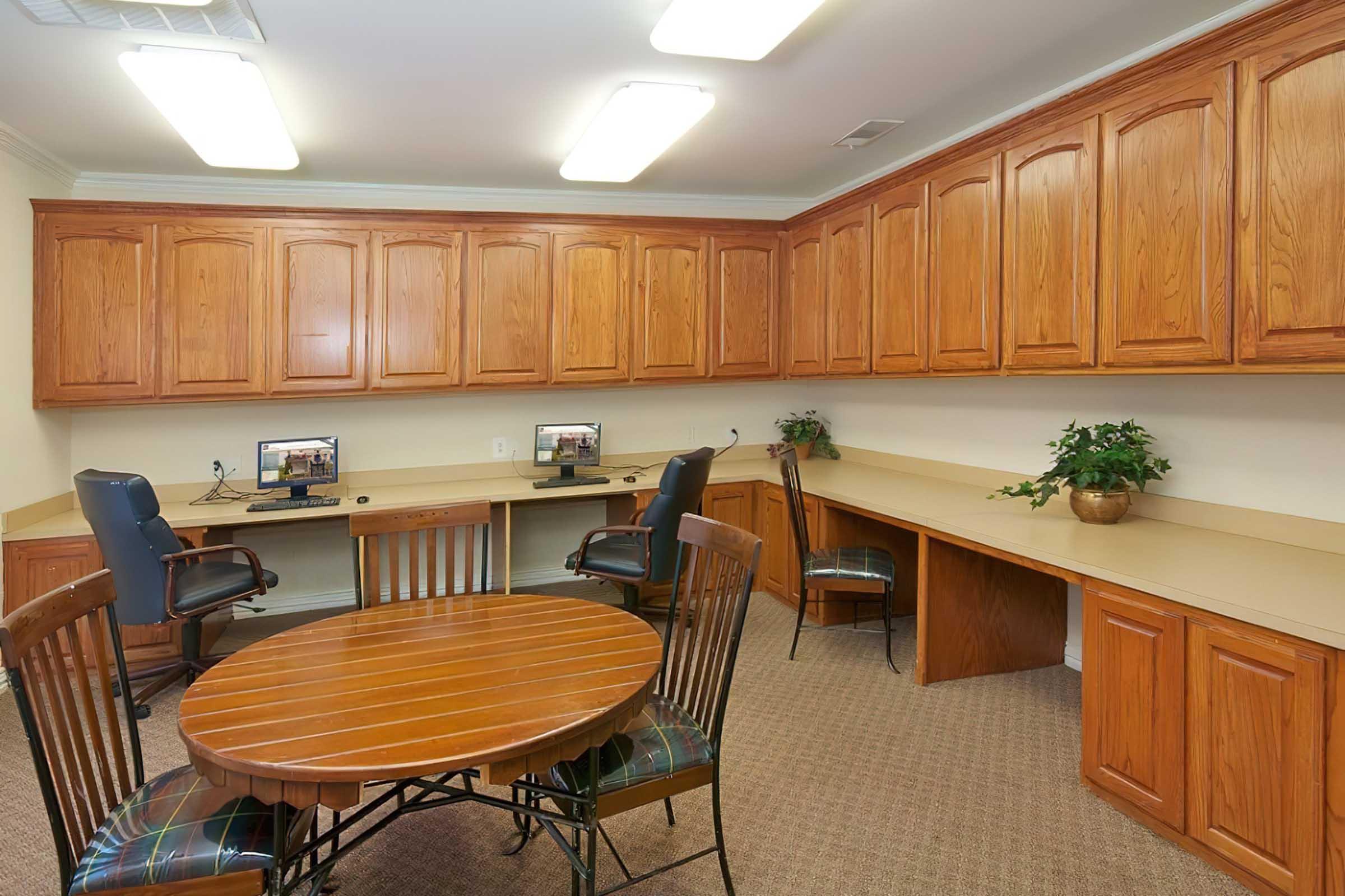 A well-lit office space featuring two computer workstations with black ergonomic chairs and a round wooden table at the center. The room has light-colored walls and wooden cabinetry above the countertops. A small potted plant adds a touch of greenery to the workspace, which has a carpeted floor.