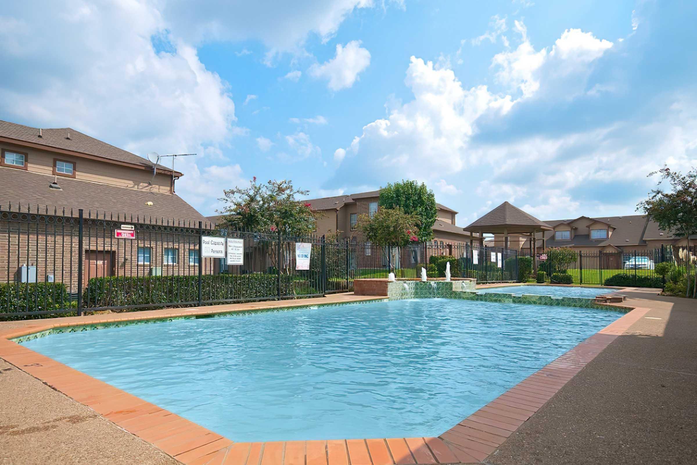 A clear swimming pool surrounded by a brick patio and decorative landscaping. Lush green bushes and trees frame the pool area, with some signage visible around the space. The sky is partly cloudy, creating a bright and inviting atmosphere in the residential area.