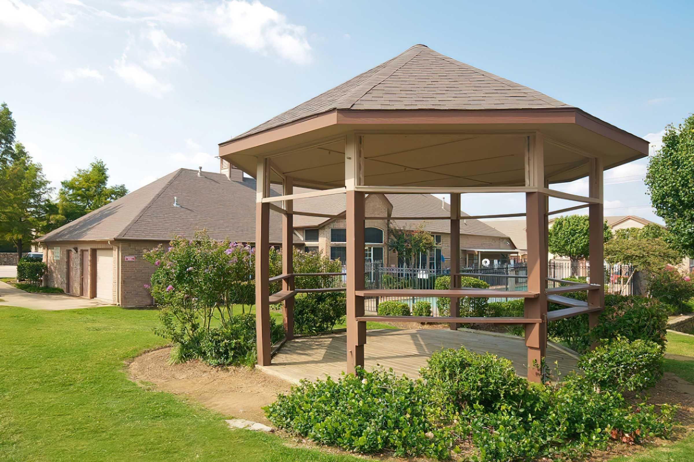 A wooden gazebo with a brown frame is situated in a landscaped area, surrounded by greenery and flowering bushes. In the background, a residential building and a swimming pool are partially visible under a clear blue sky, creating a serene outdoor setting.