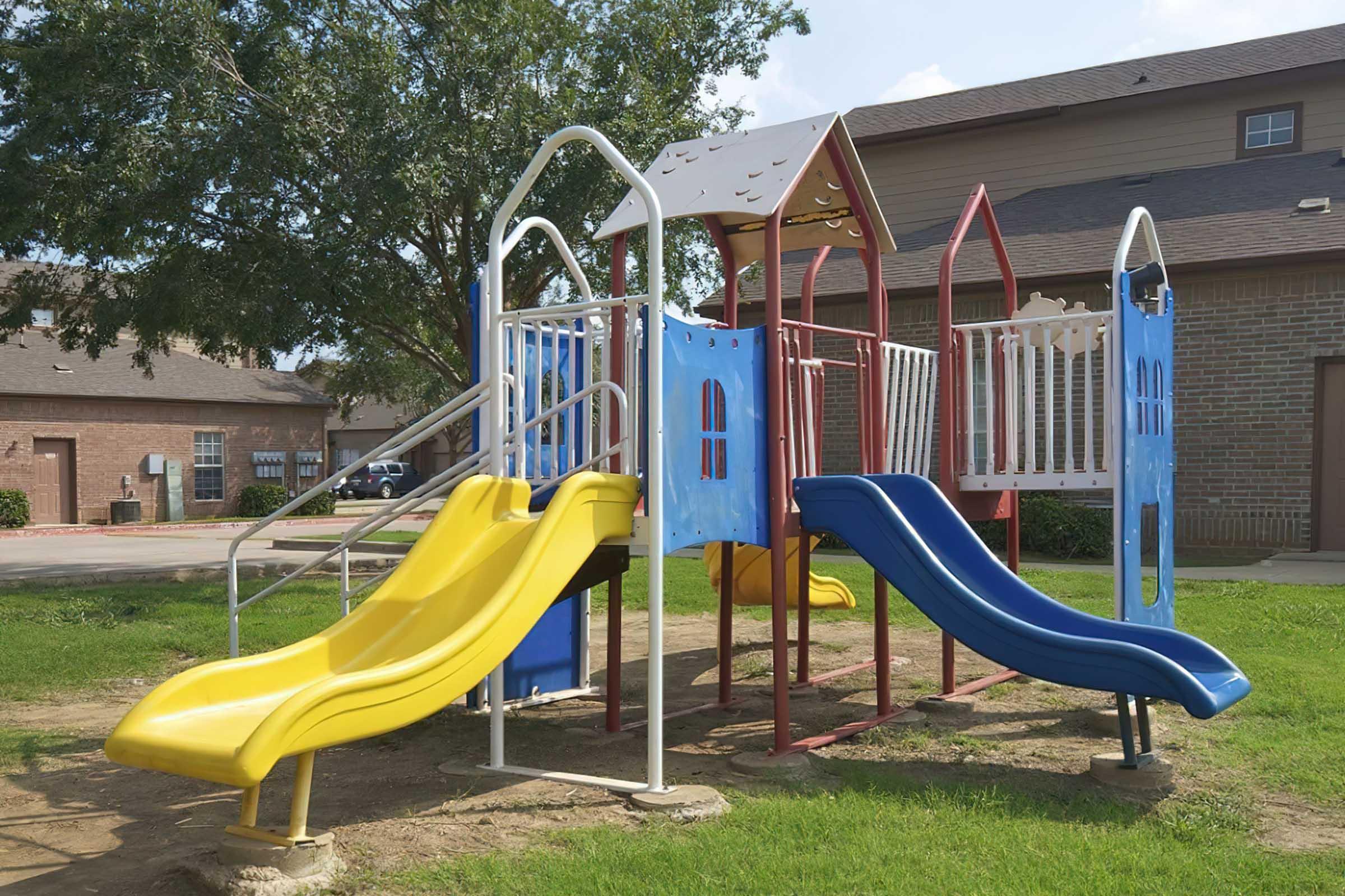 A colorful playground structure featuring two slides—one yellow and one blue—alongside climbing features. The playground is situated in a grassy area with nearby buildings in the background and trees providing shade.