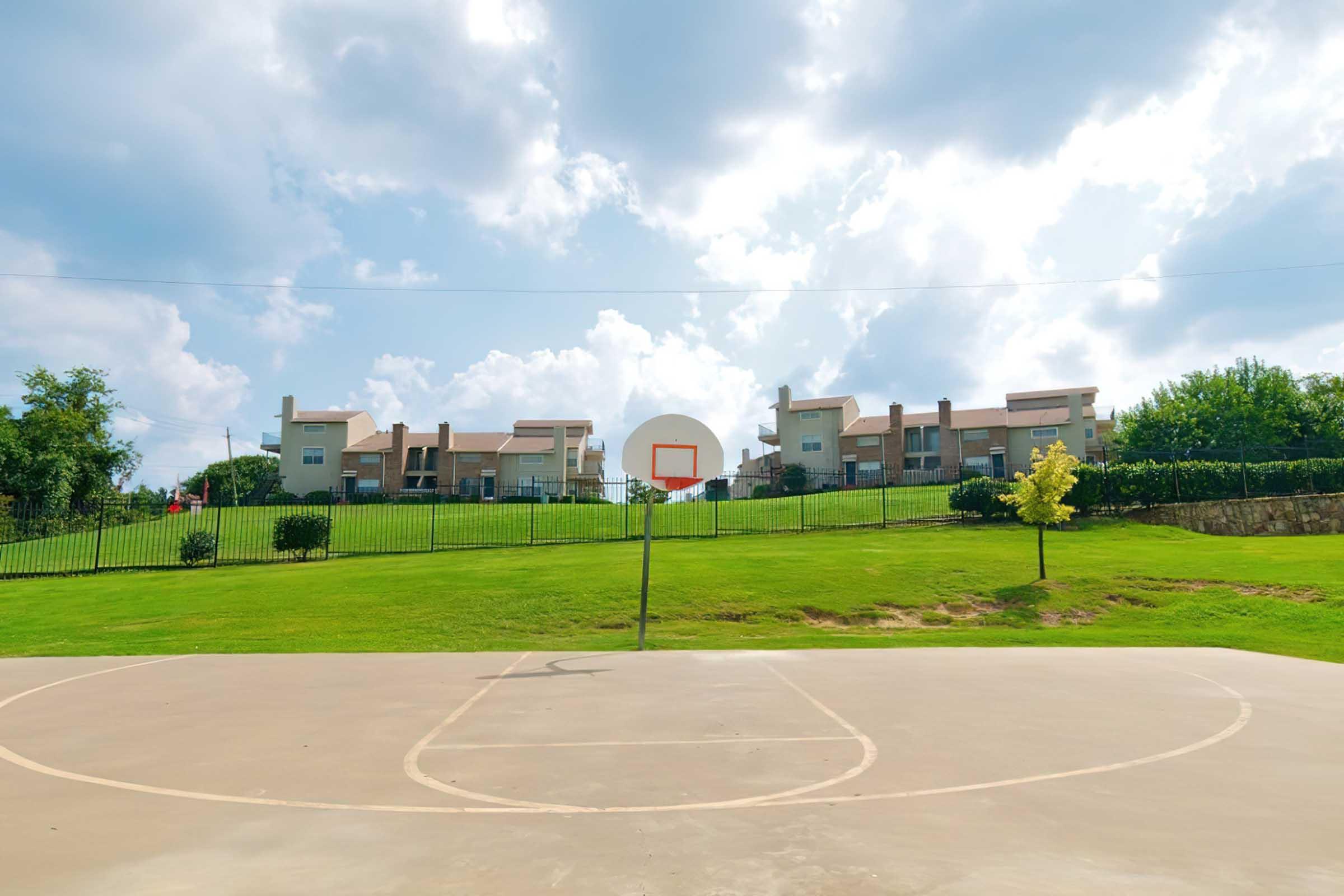 A basketball court in the foreground with a hoop and a smooth surface. In the background, there are several multi-story apartment buildings on a grassy hill, surrounded by trees and a fence under a partly cloudy sky.