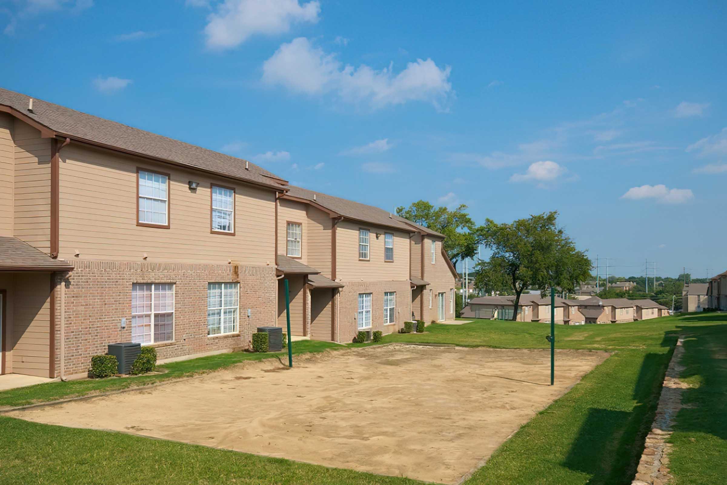 A row of beige two-story apartment buildings with large windows, set on a well-maintained green lawn. A sandy area is visible in front, with a basketball hoop. The sky is clear with a few white clouds, suggesting a sunny day.