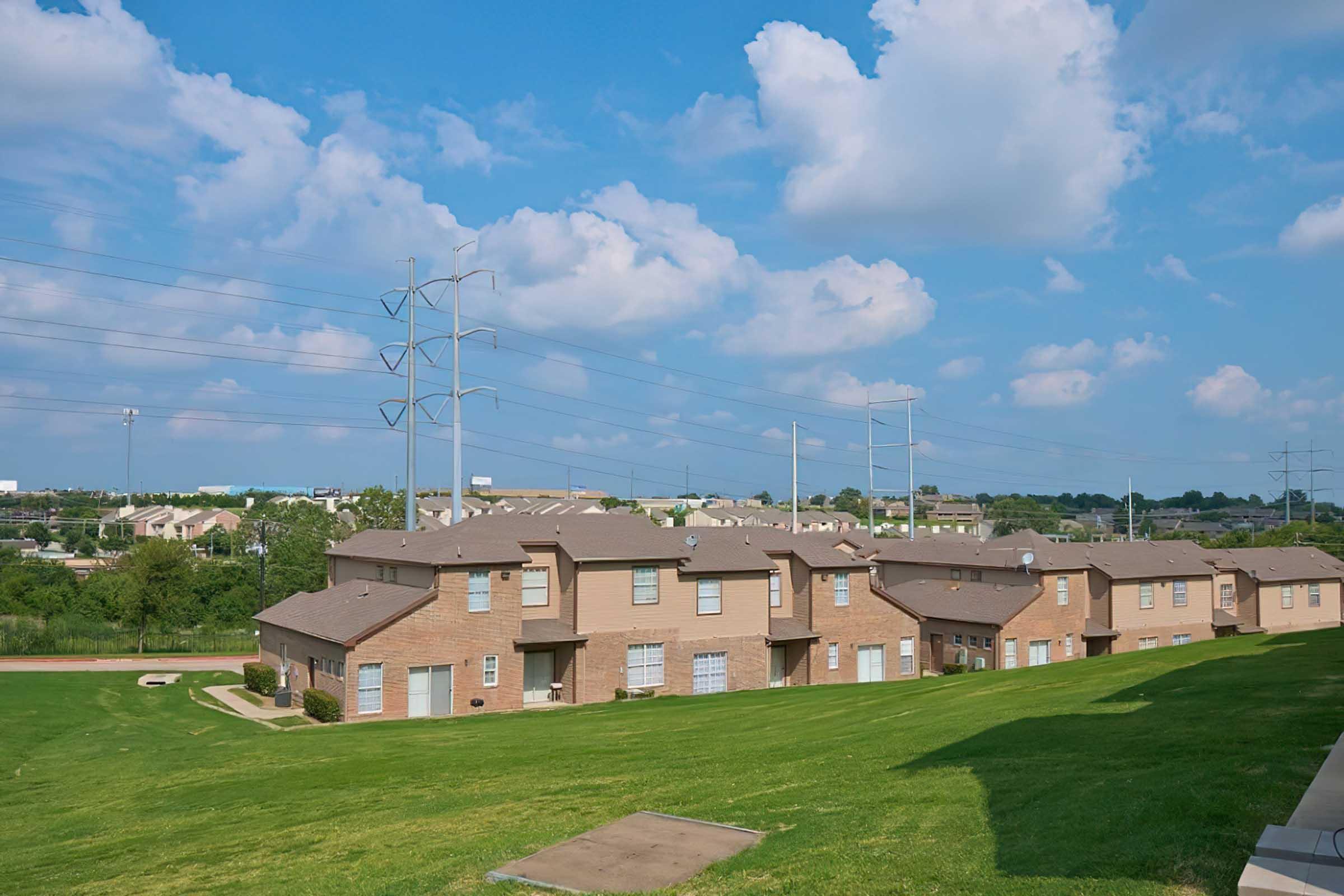 A view of a residential area featuring low-rise brick apartment buildings set on a grassy hillside. The background includes several tall utility poles and scattered clouds against a blue sky, with more buildings visible in the distance.